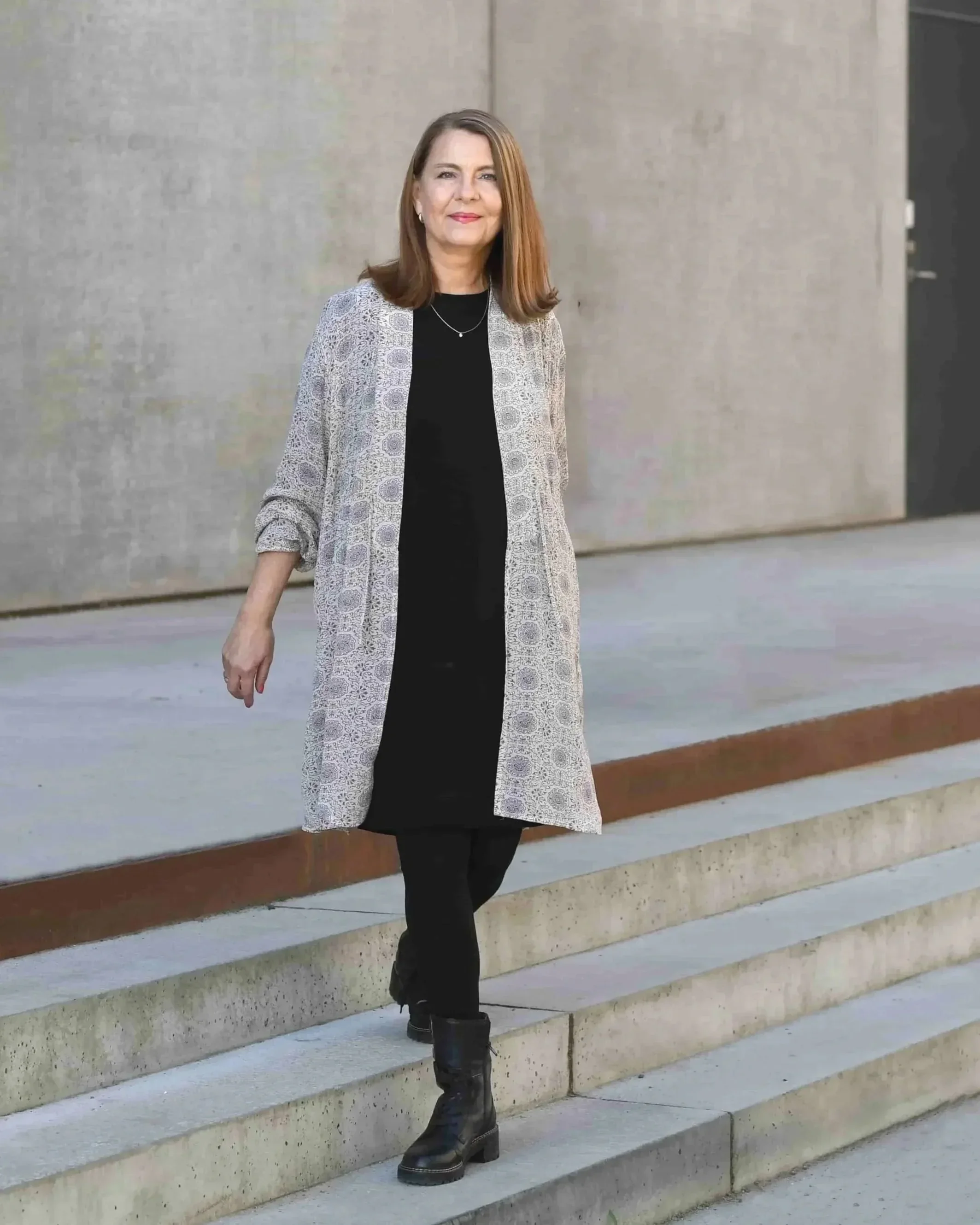 Portrætbillede af Designcaoch founder Angela  Lund , der går ned ad udendørs trin, iført sort tøj, støvler og en lang, lysegrå jakke med mønster, foran en betongvæg.