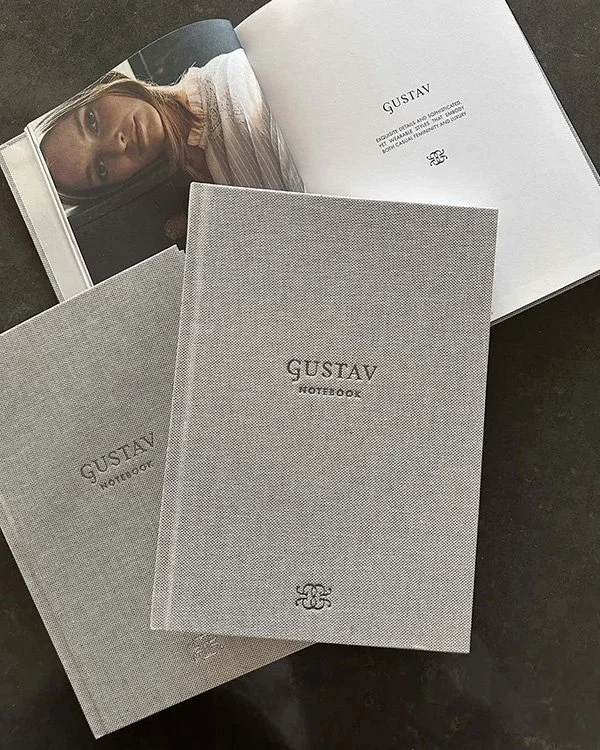 To grafiske notesbøger desiget af Angela Lund/designcoach for Guastav Denmark med 'GUSTAV NOTEBOOK' skrevet på forsiden og en åben bog med et billede af en kvinde.
