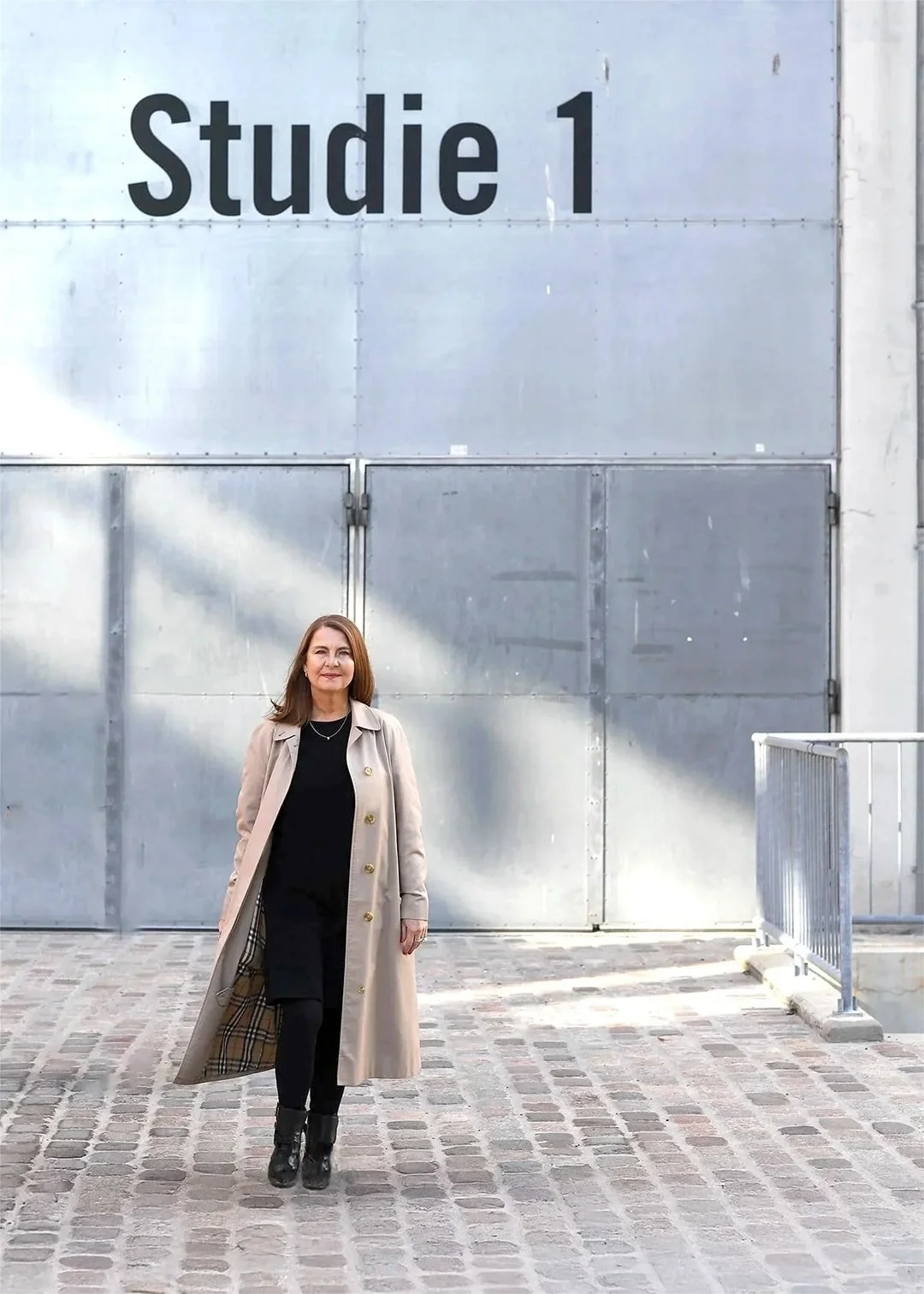 Designcoach, webdesigner og brandarkitekt Angela Lund fotograferet ude, foran en stor metalport i Filmbyen Aarhus, Danmark, med skiltet 'Studie 1', iført en beige lang frakke, sort tøj og støvler, ved en grusbelagt gård.