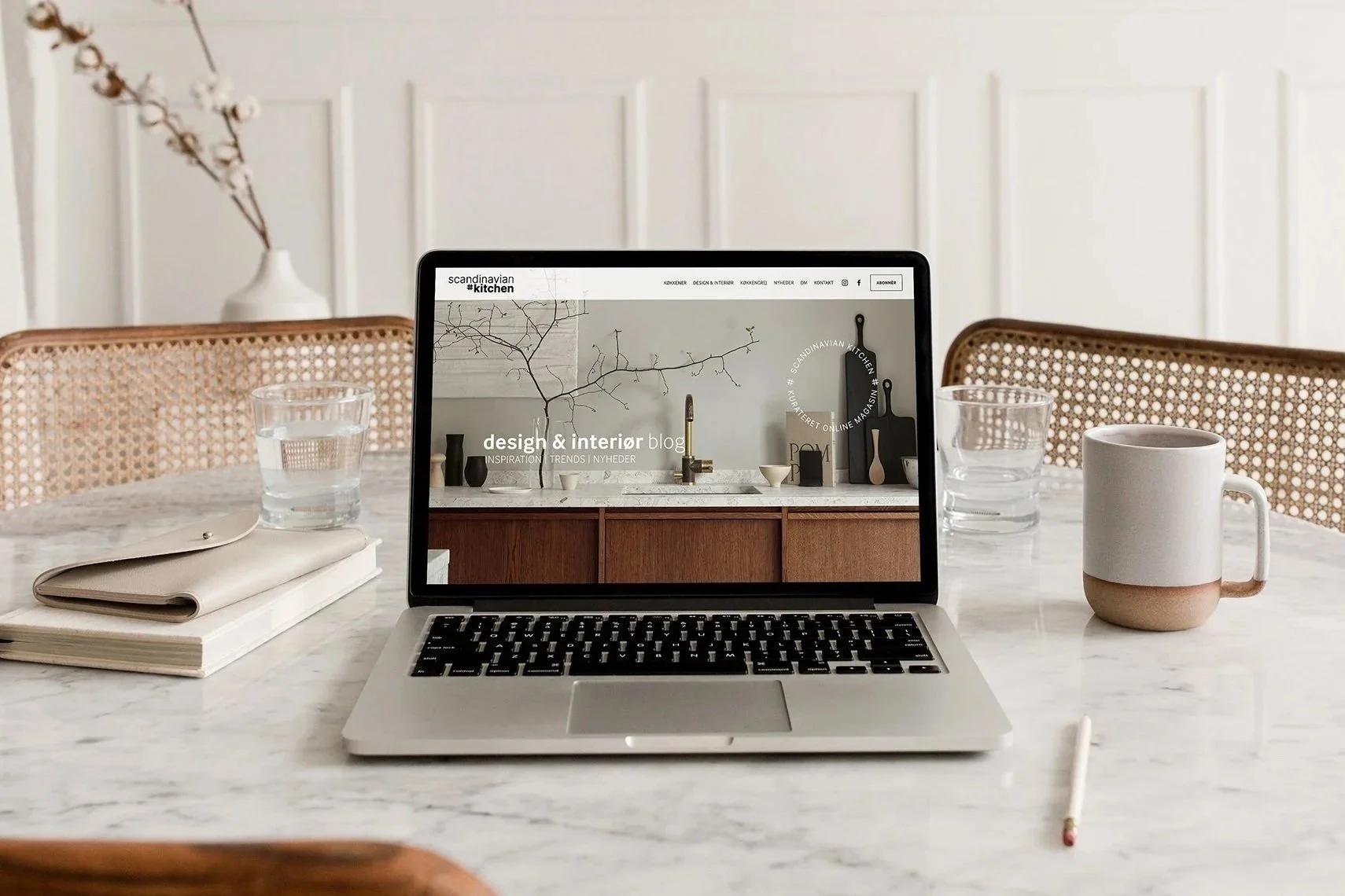 En laptop, der viser en hjemmeside designet af designcoach.dk, på et marmorbord, to glas med vand, en tallerken, en notesbog, en pen, en kop, og to stole  mod en lys væg.