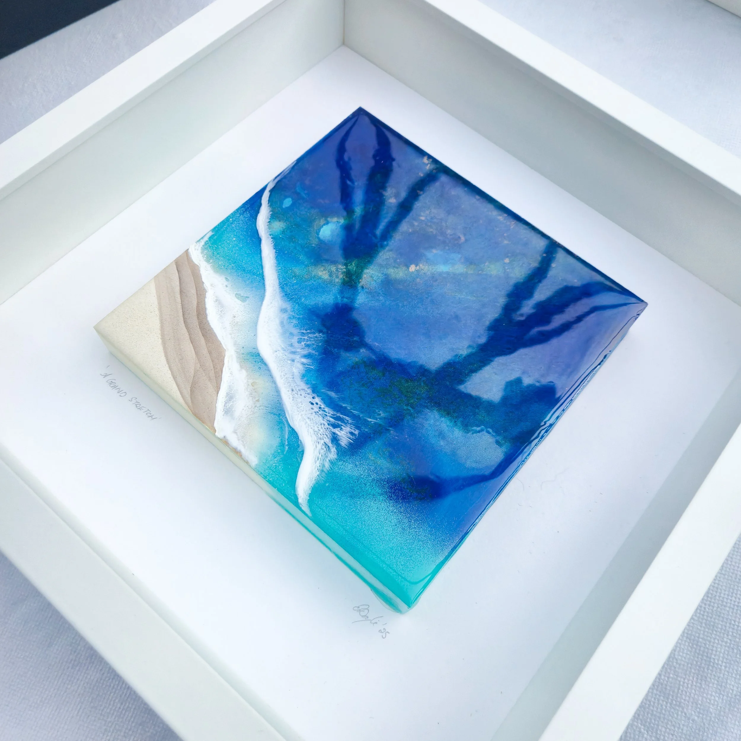 Framed resin artwork with deep blue ocean water and turquoise tones meeting sandy shore, displayed in white shadow box frame.