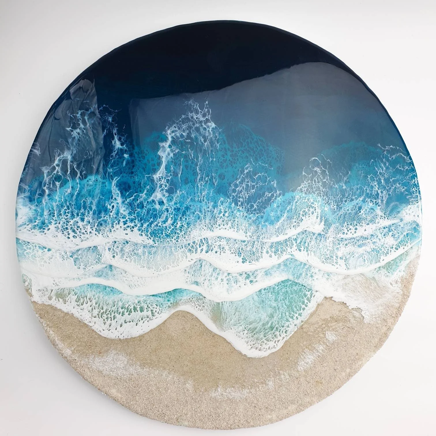 Circular art piece depicting ocean waves crashing onto sandy beach with foam and blue water, on a white background.