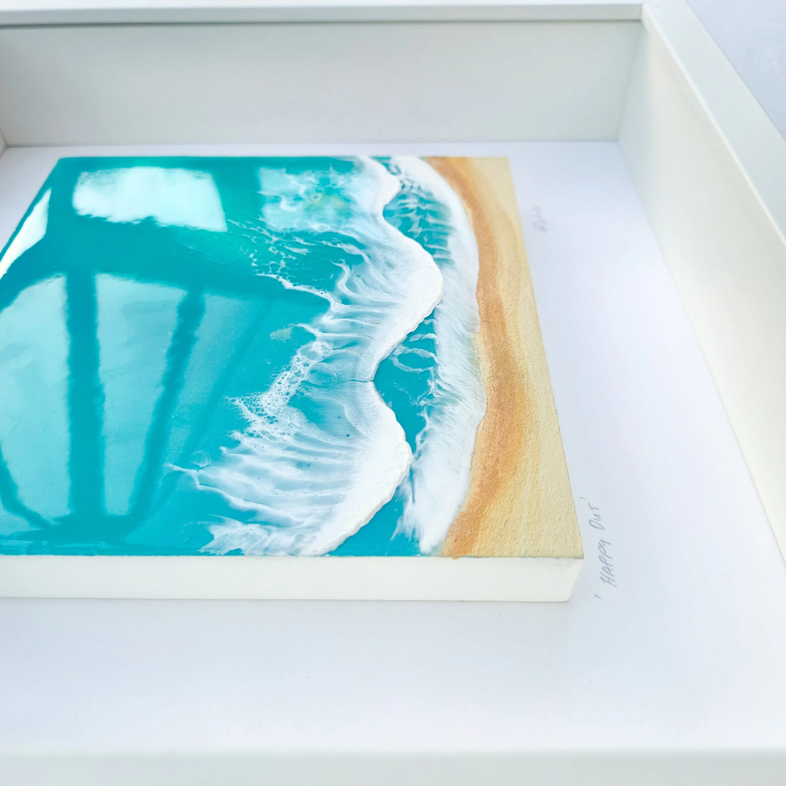 Framed resin artwork with turquoise ocean waves and sandy beach, displayed in white shadow box with glass-like finish.
