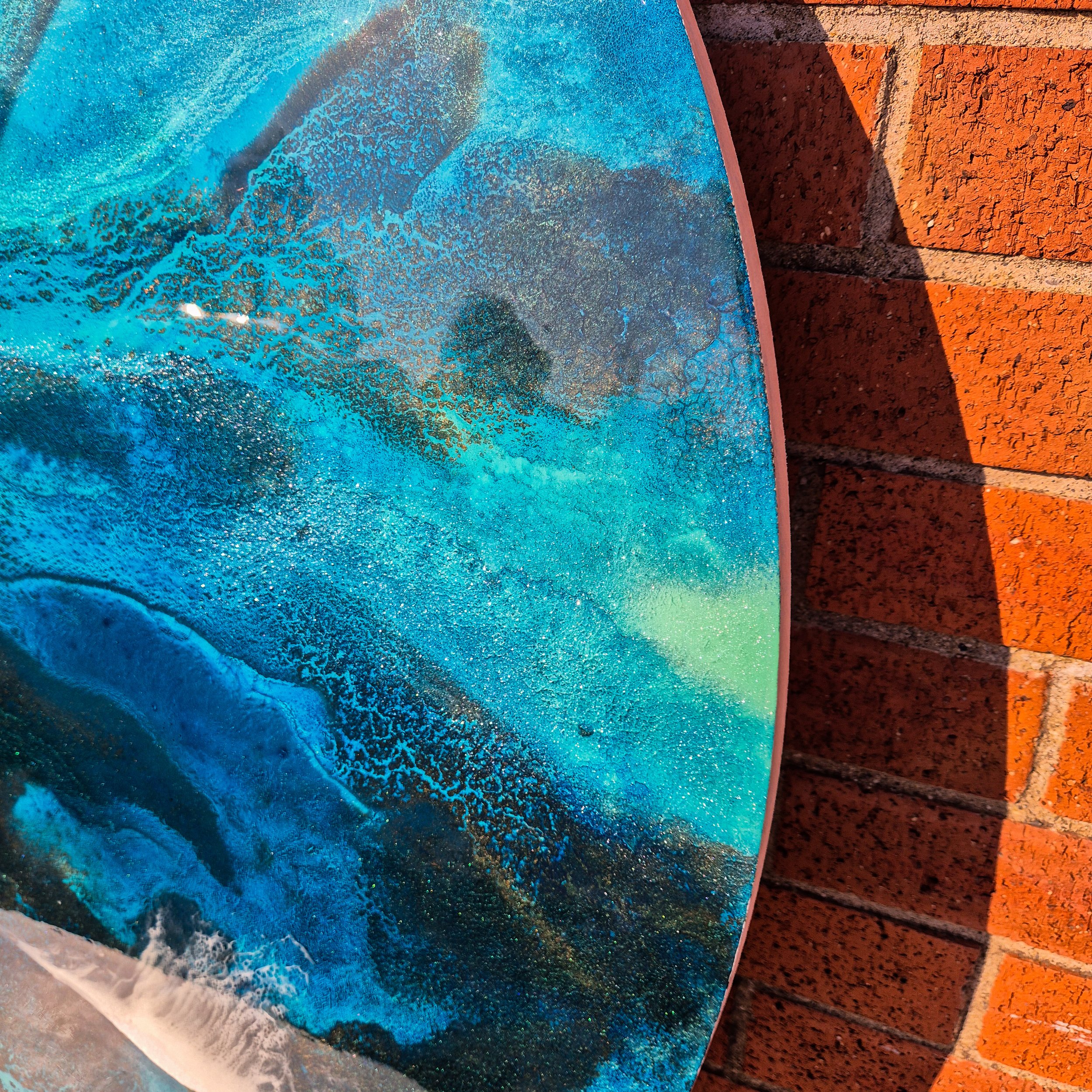 Large round resin artwork with swirling blue ocean textures and sandy shoreline, featuring a high-gloss finish on wood panel.