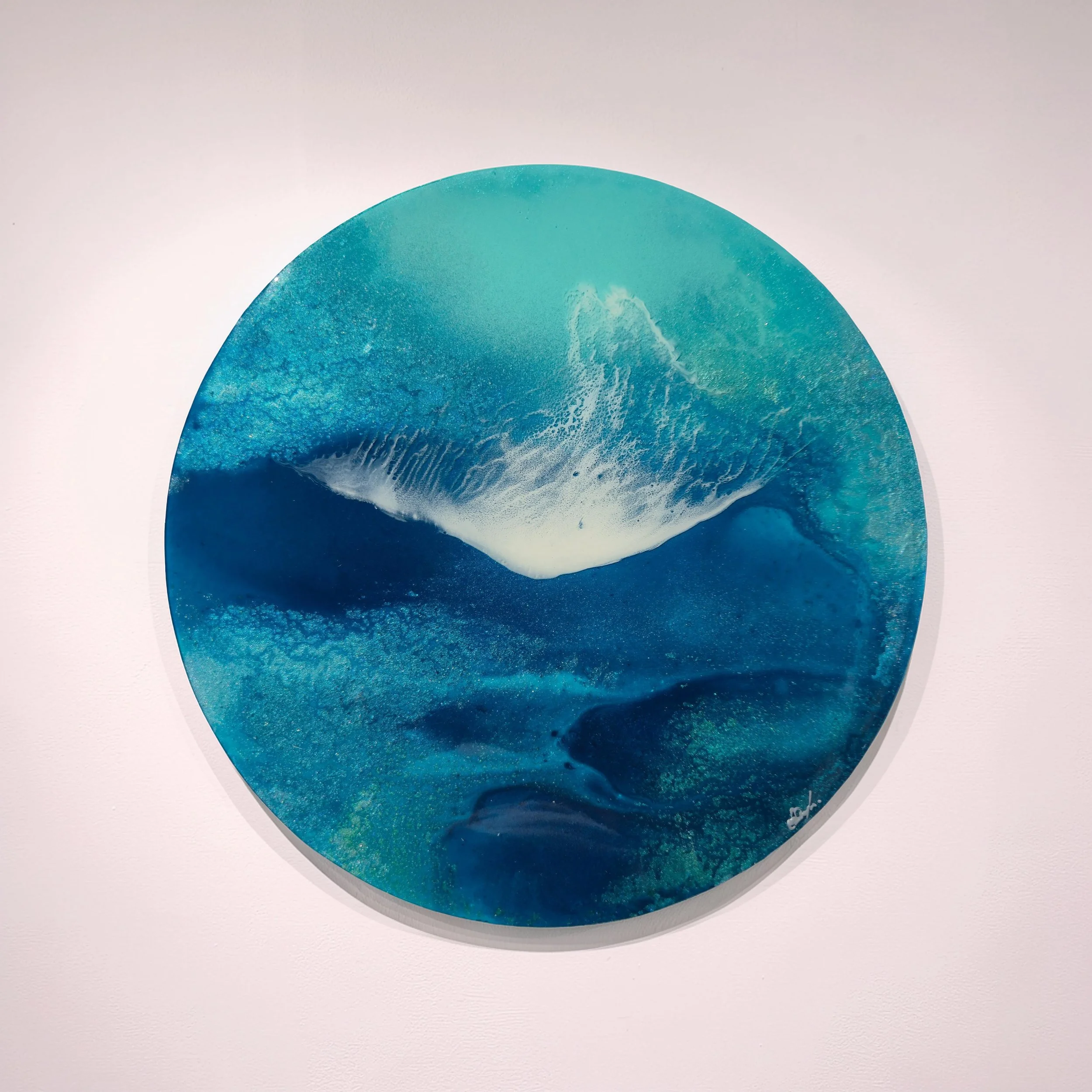 Easy Tide round resin artwork on a pale wall. Teal water and a soft white wave seen from above.