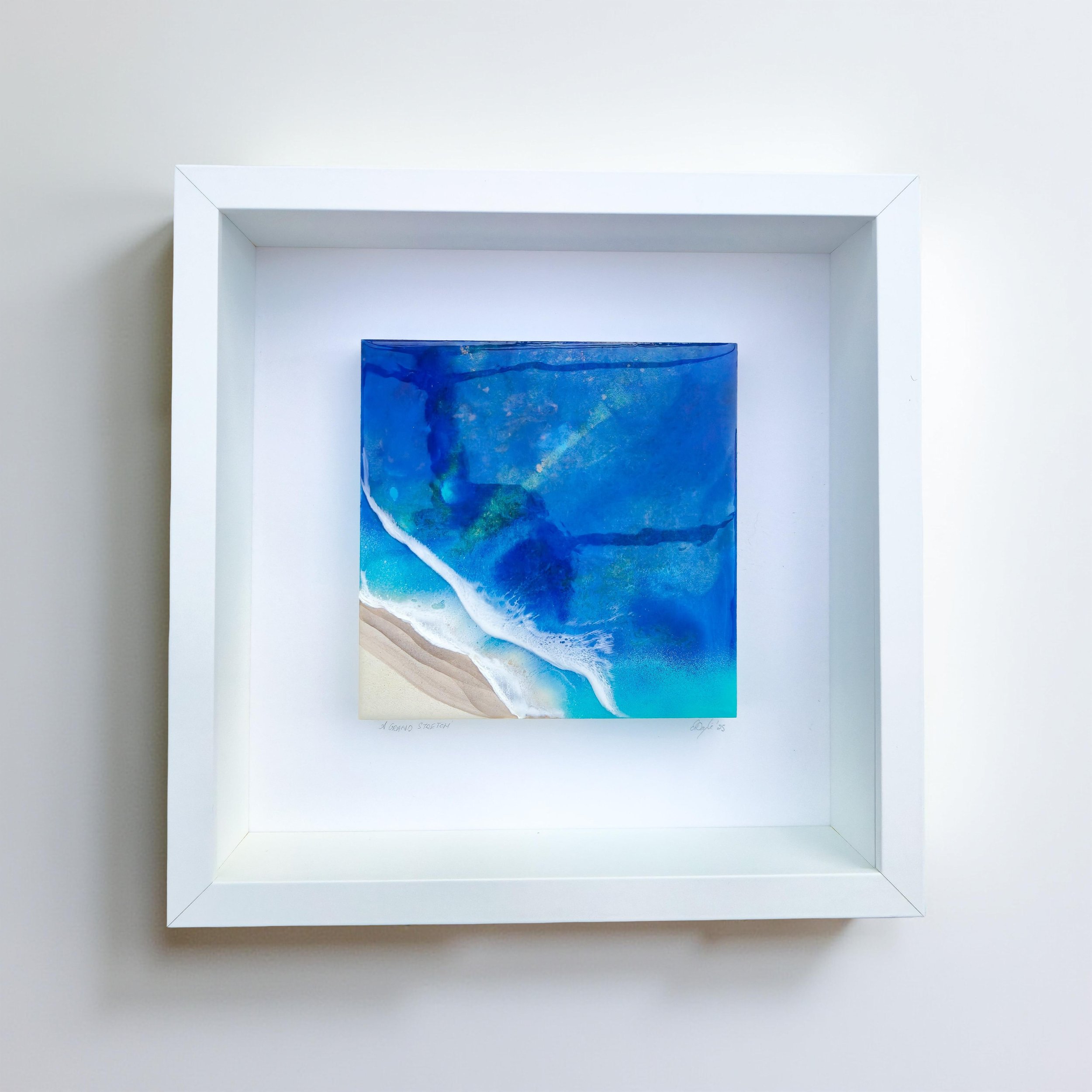 Framed resin artwork with deep blue ocean water and turquoise tones meeting sandy shore, displayed in white shadow box frame.