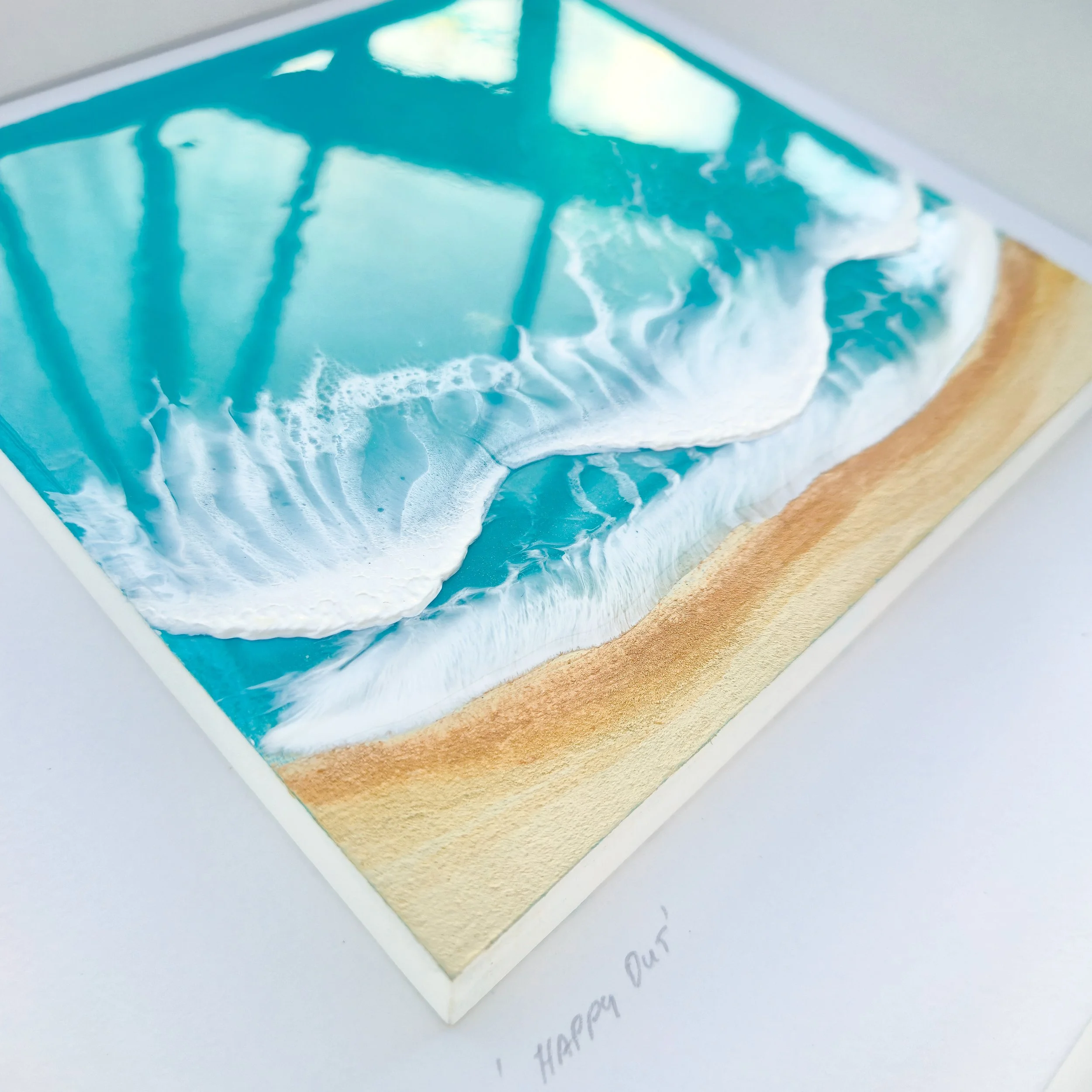Framed resin artwork with turquoise ocean waves and sandy beach, displayed in white shadow box with glass-like finish.