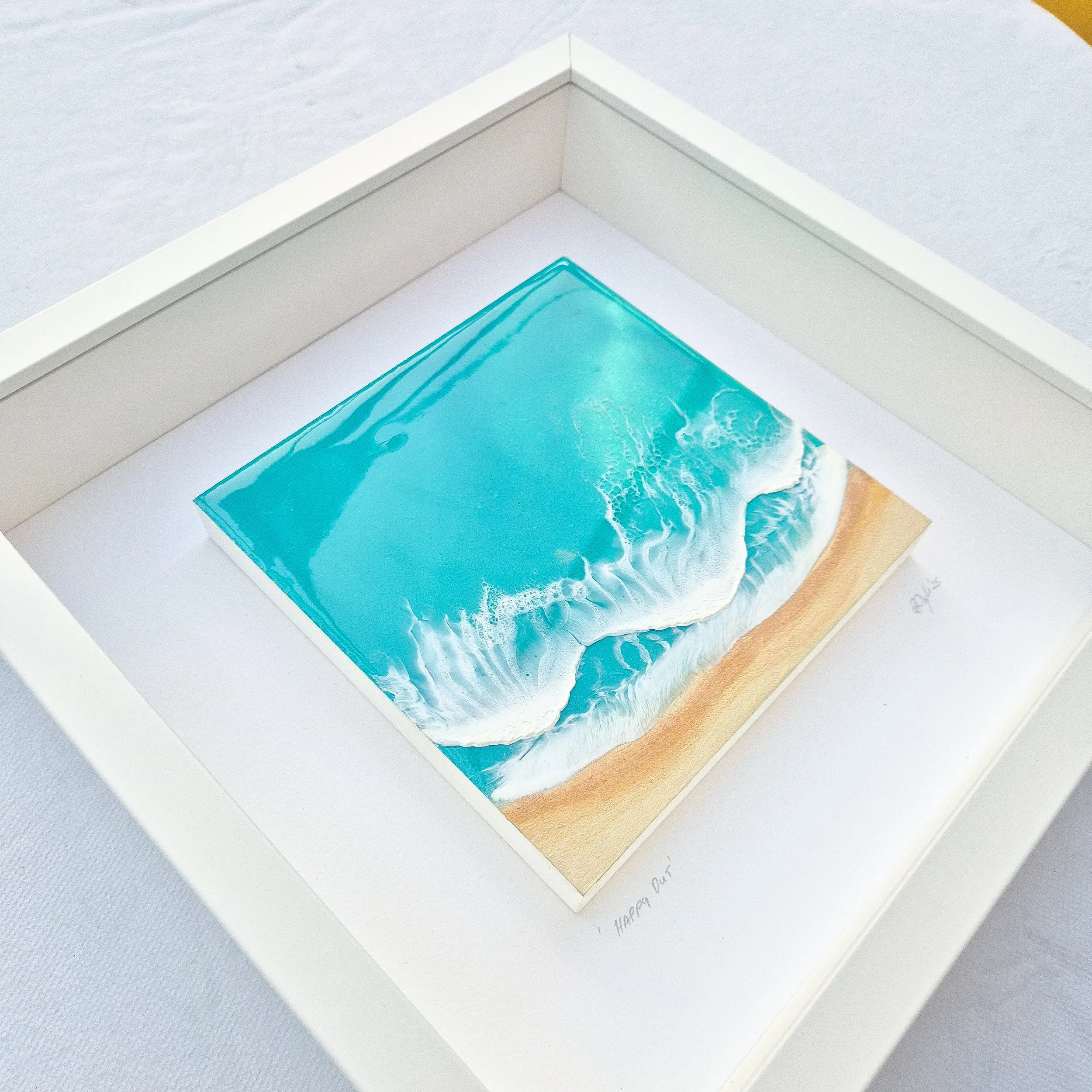 Framed resin artwork with turquoise ocean waves and sandy beach, displayed in white shadow box with glass-like finish.