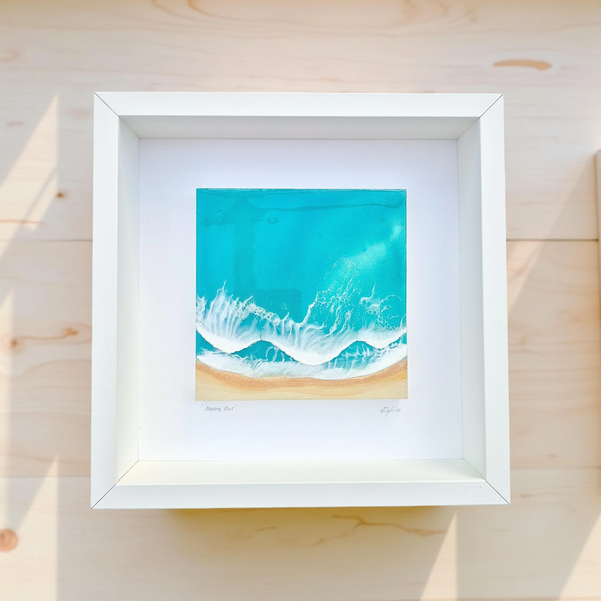 Framed resin artwork with turquoise ocean waves and sandy beach, displayed in white shadow box with glass-like finish.