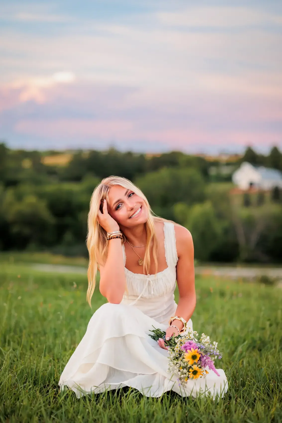 Senior girl wearing a white dress sitting on grass, holding a bouquet of flowers, smiling at the camera with a scenic countryside background and colorful sky. Photo by Wendy Sorensen, a Des Moines area senior photographer.