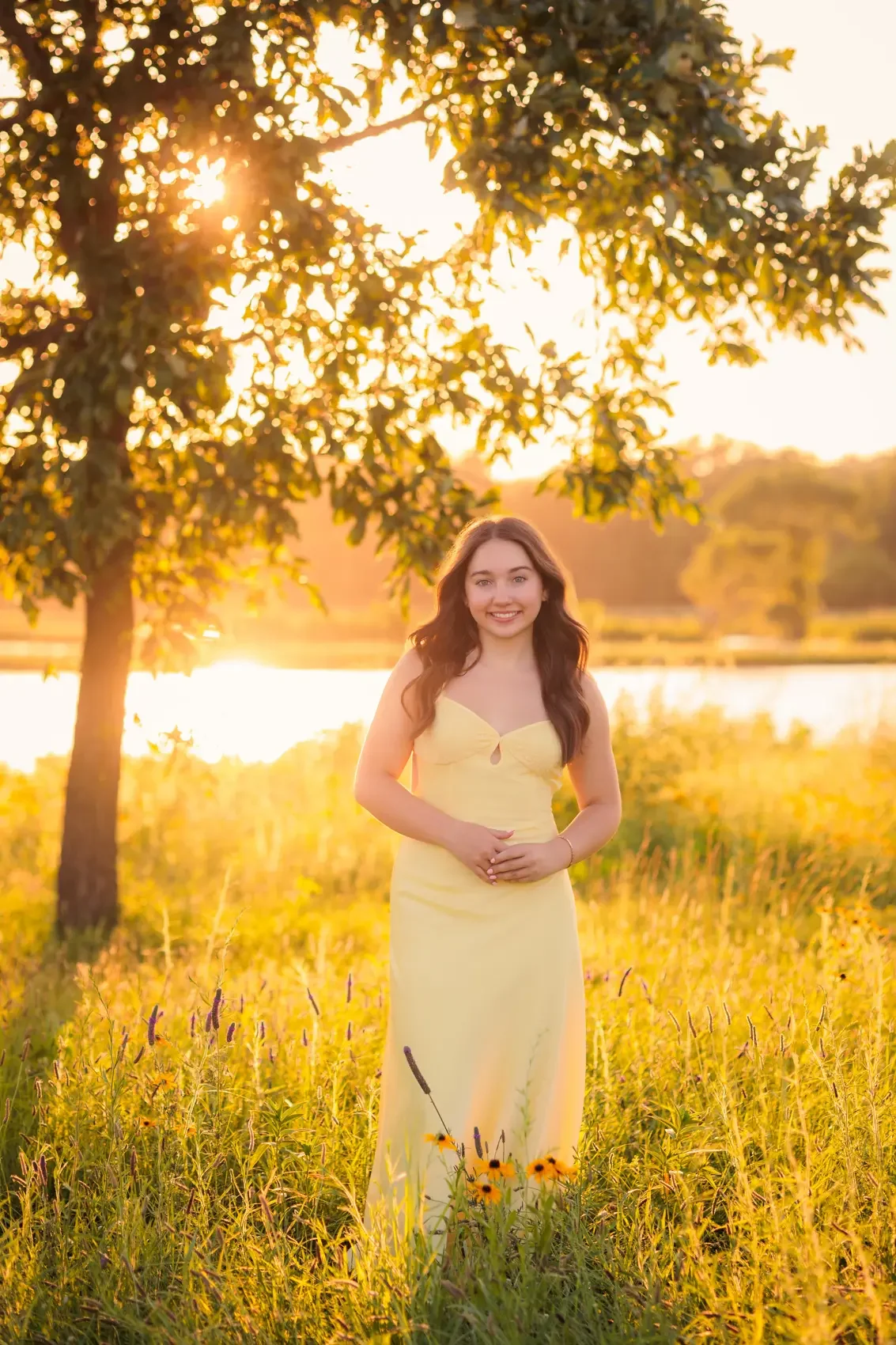 Young woman in a yellow dress standing in a field of yellow flowers during sunset, with a tree and a river in the background.