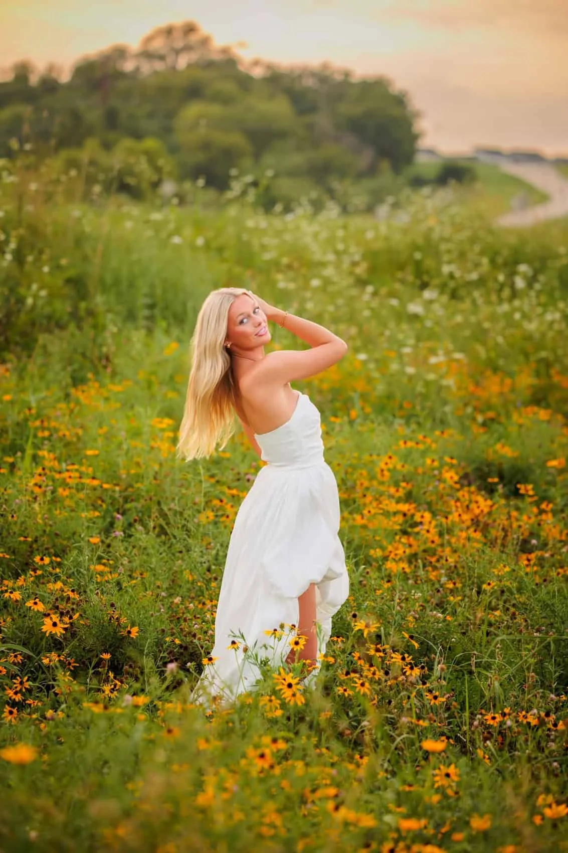 Senior girl in a white dress standing in a field of yellow and orange flowers with green foliage, during sunset. Photo by Wendy Sorensen, a Des Moines senior photographer.