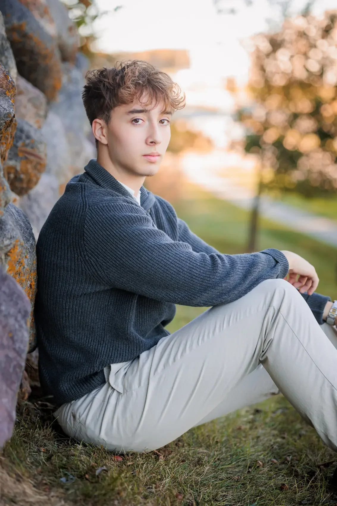 A young man sitting on the grass beside a stone wall outdoors during sunset, wearing a navy blue sweater and light-colored pants.