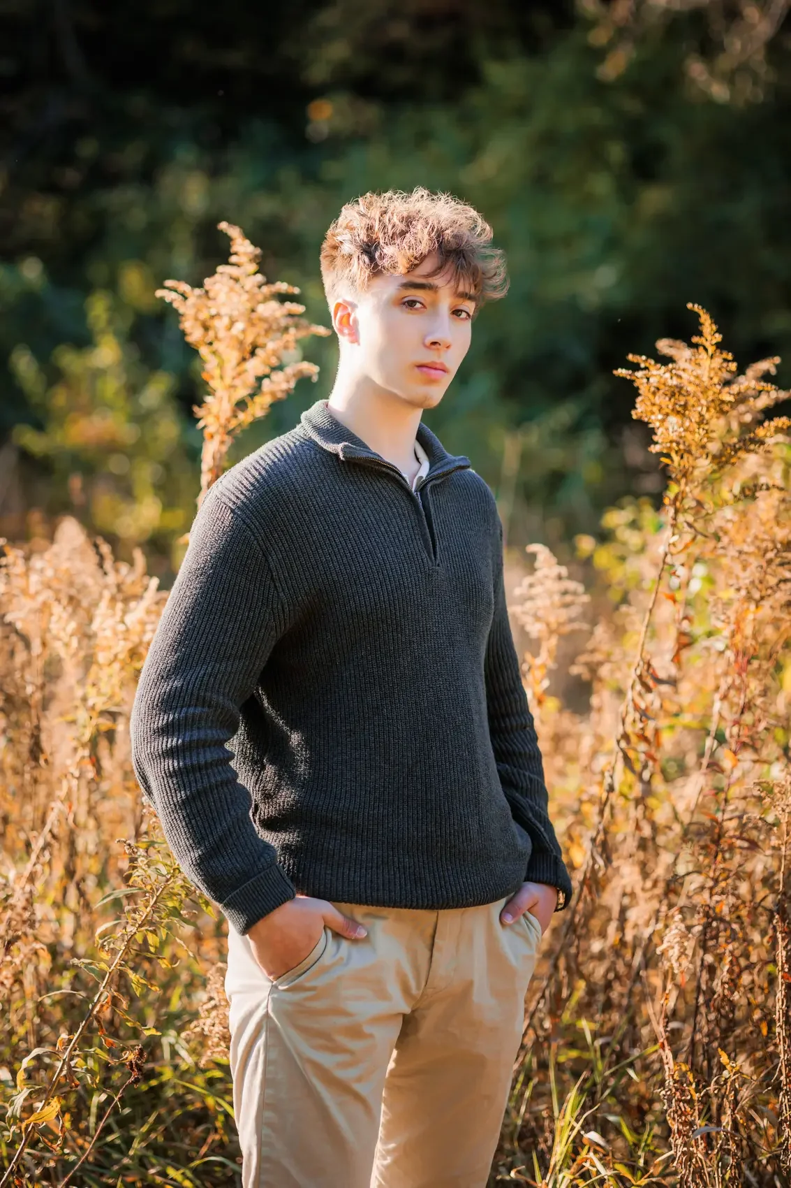 A young man with curly hair stands outdoors in a field of golden plants during autumn, wearing a black sweater and beige pants, with hands in his pockets, looking at the camera.