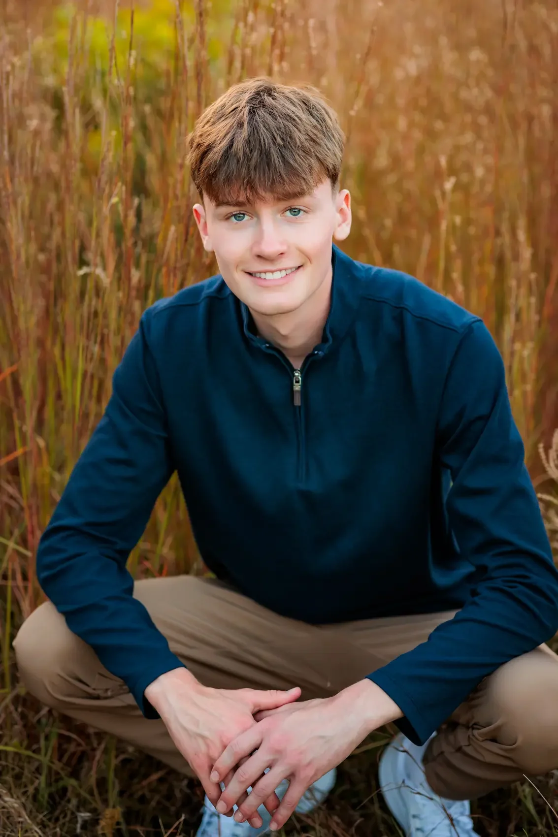 Senior boy with light brown hair and blue eyes, wearing a navy zipped-up jacket and khaki pants, squatting outdoors among tall grasses with golden-brown hues, smiling at the camera.