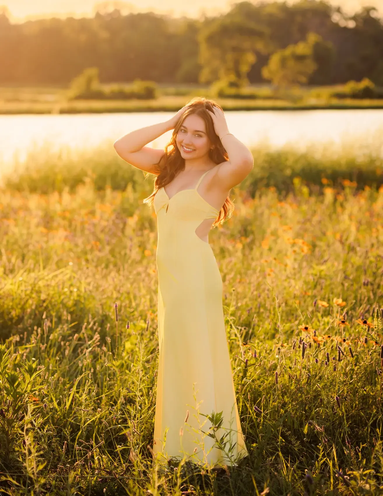 Senior girl in a yellow dress standing in a field of wildflowers near a body of water at sunset at Water Works Park in Des Moines, Iowa for a senior photo session.