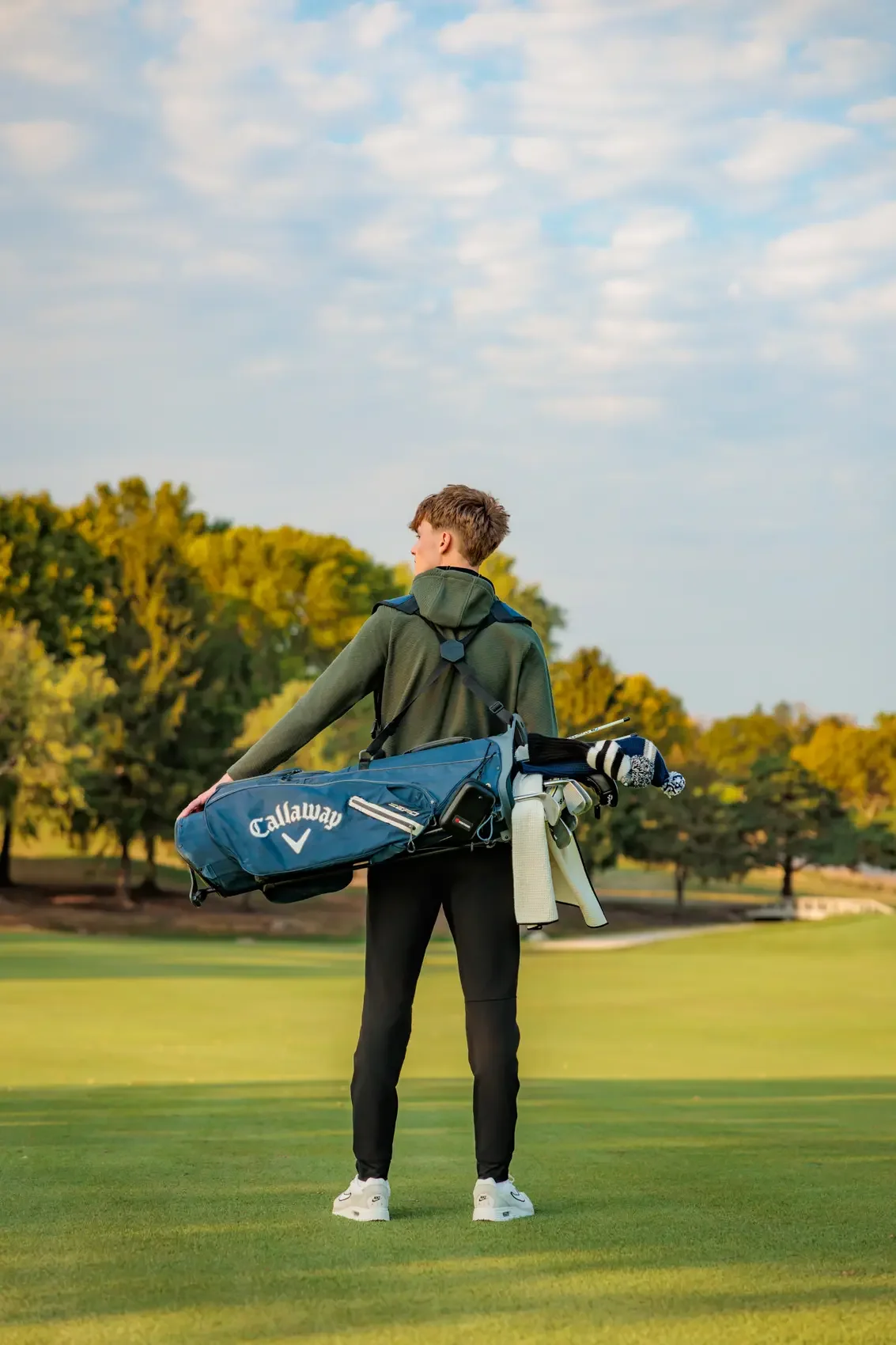 Senior boy standing on a golf course holding a golf bag with clubs, dressed in casual sportswear, with trees and a cloudy sky in the background.