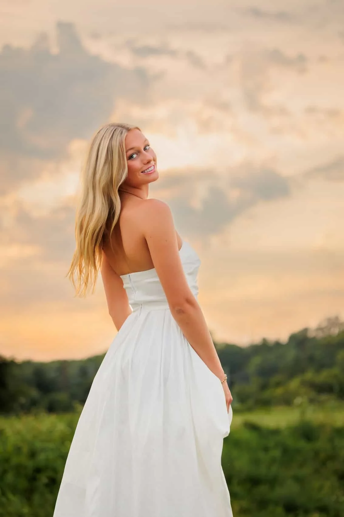 Senior girl in a white strapless dress standing outdoors during sunset, with a background of cloudy sky and greenery.