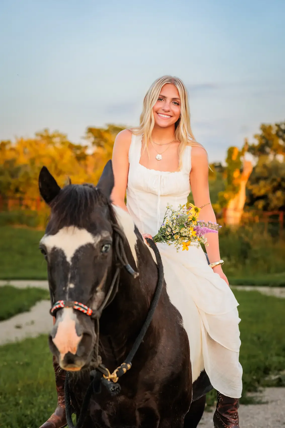 High school senior girl in a white dress is sitting on a black and white horse, holding a bouquet of wildflowers. She is smiling outdoors during sunset with autumn trees in the background.
