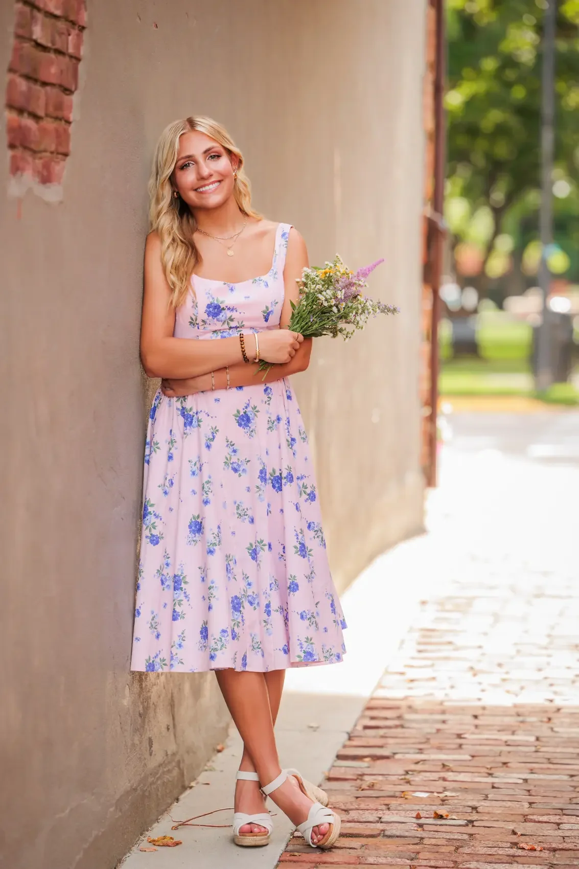Senior girl standing against a wall outdoors, holding a bouquet of wildflowers, wearing a light purple floral dress and white wedge sandals, smiling at the camera.