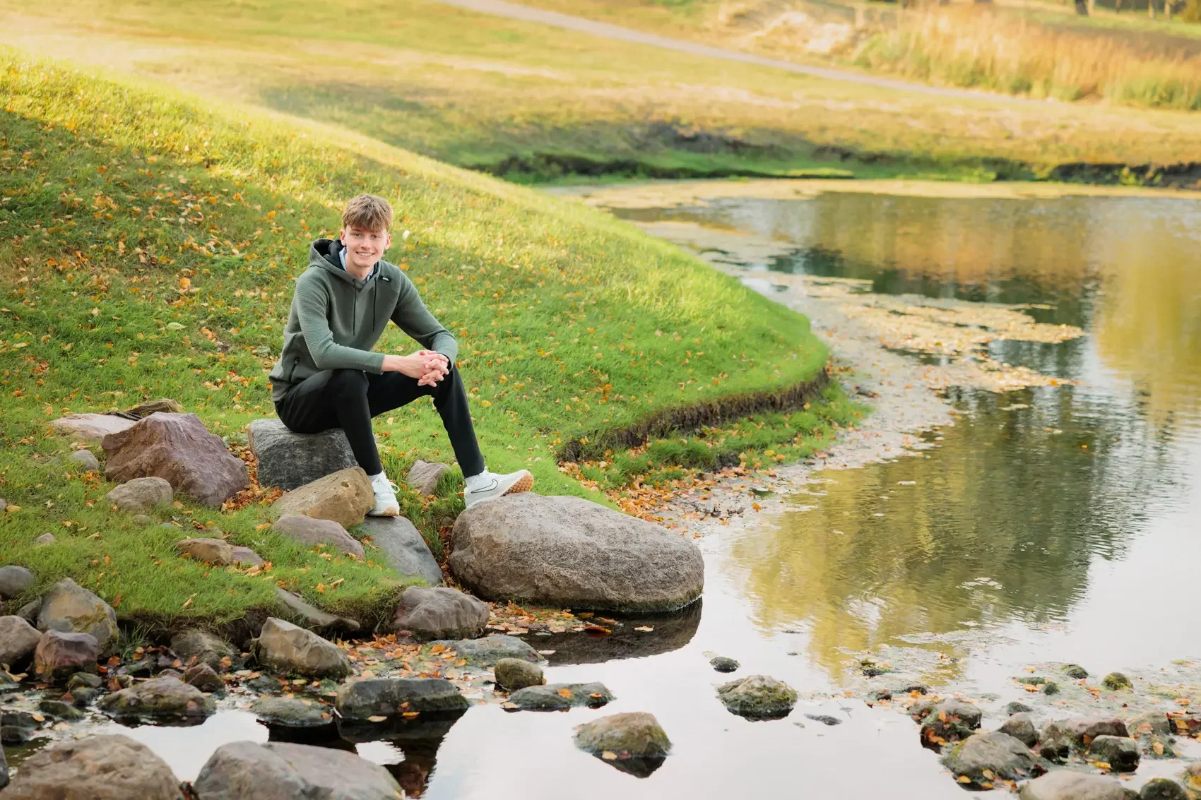 Senior boy sits on rocks by a lake surrounded by green grass and trees with fall foliage at Des Moines Golf & Country Club.