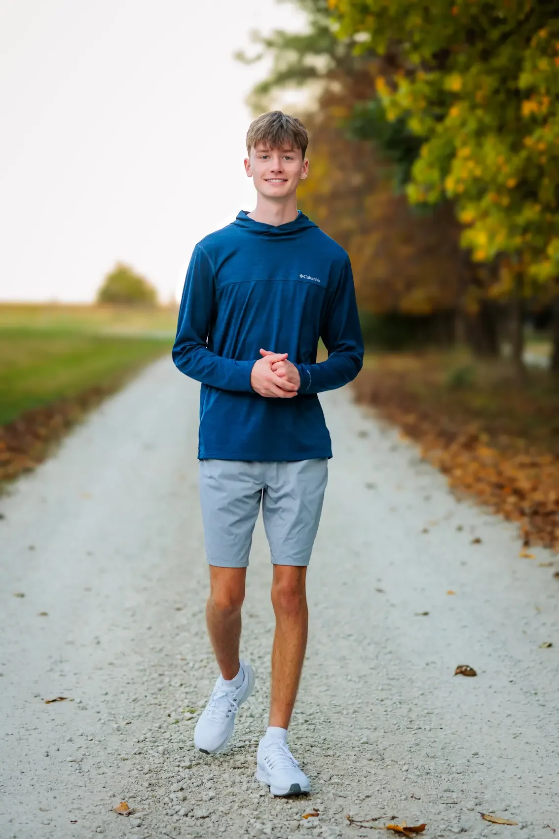 Senior boy walking outdoors on a gravel path, wearing a blue athletic hoodie, gray shorts, and white running shoes, with trees in the background.
