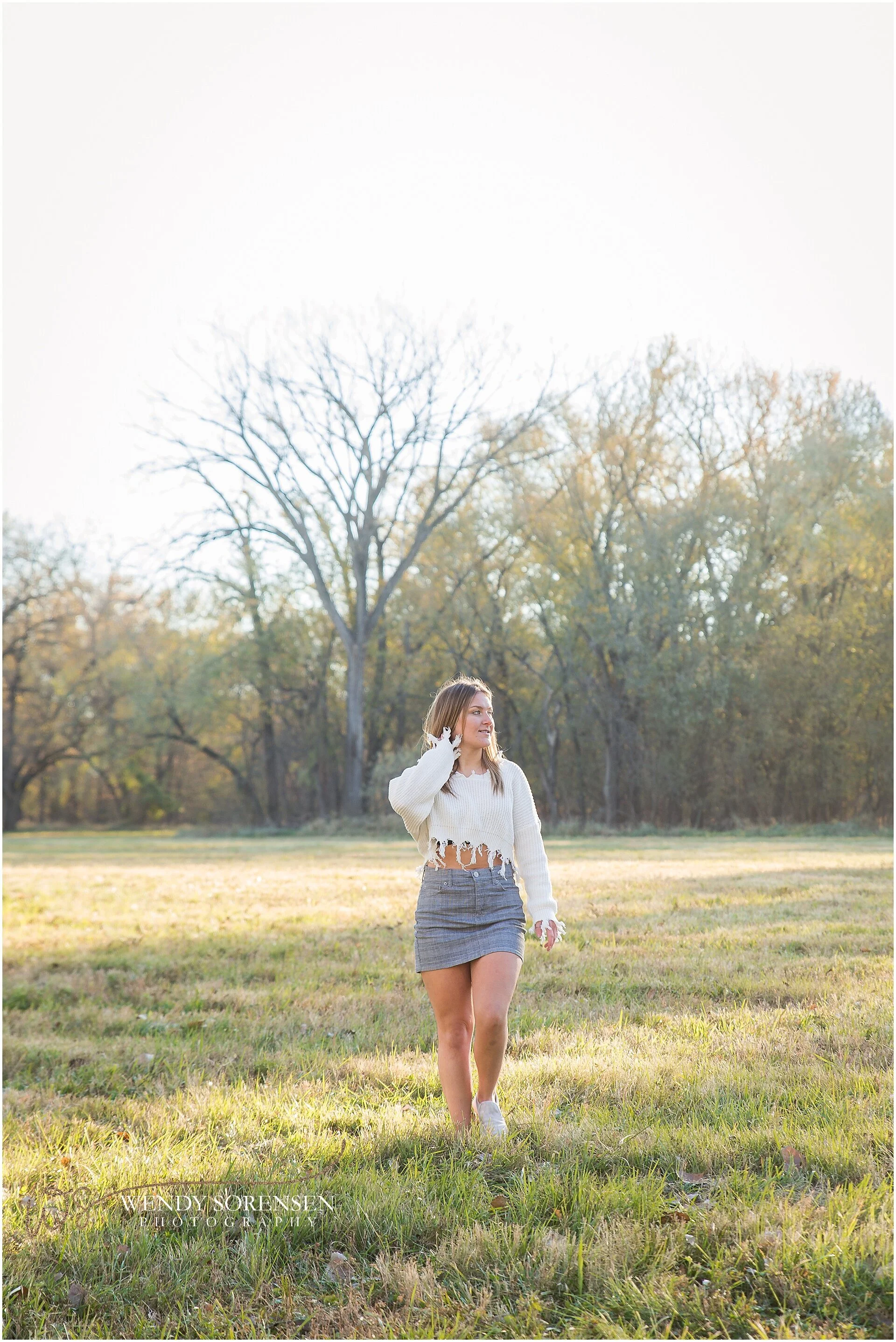 Senior girl wearing a cream sweater and gray skirt walking through the grass, looking off to the side, in Des Moines.