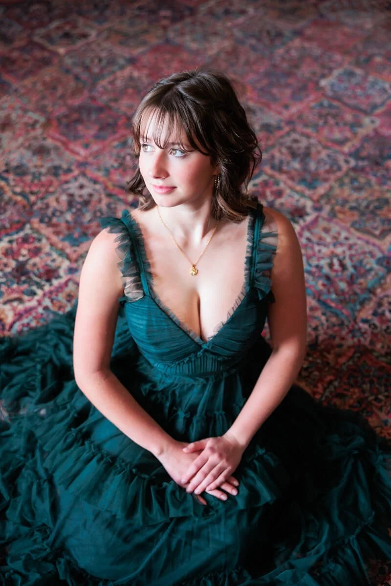 High school senior girl wearing ruffled green prom dress, sitting on a beautiful pattered rug.