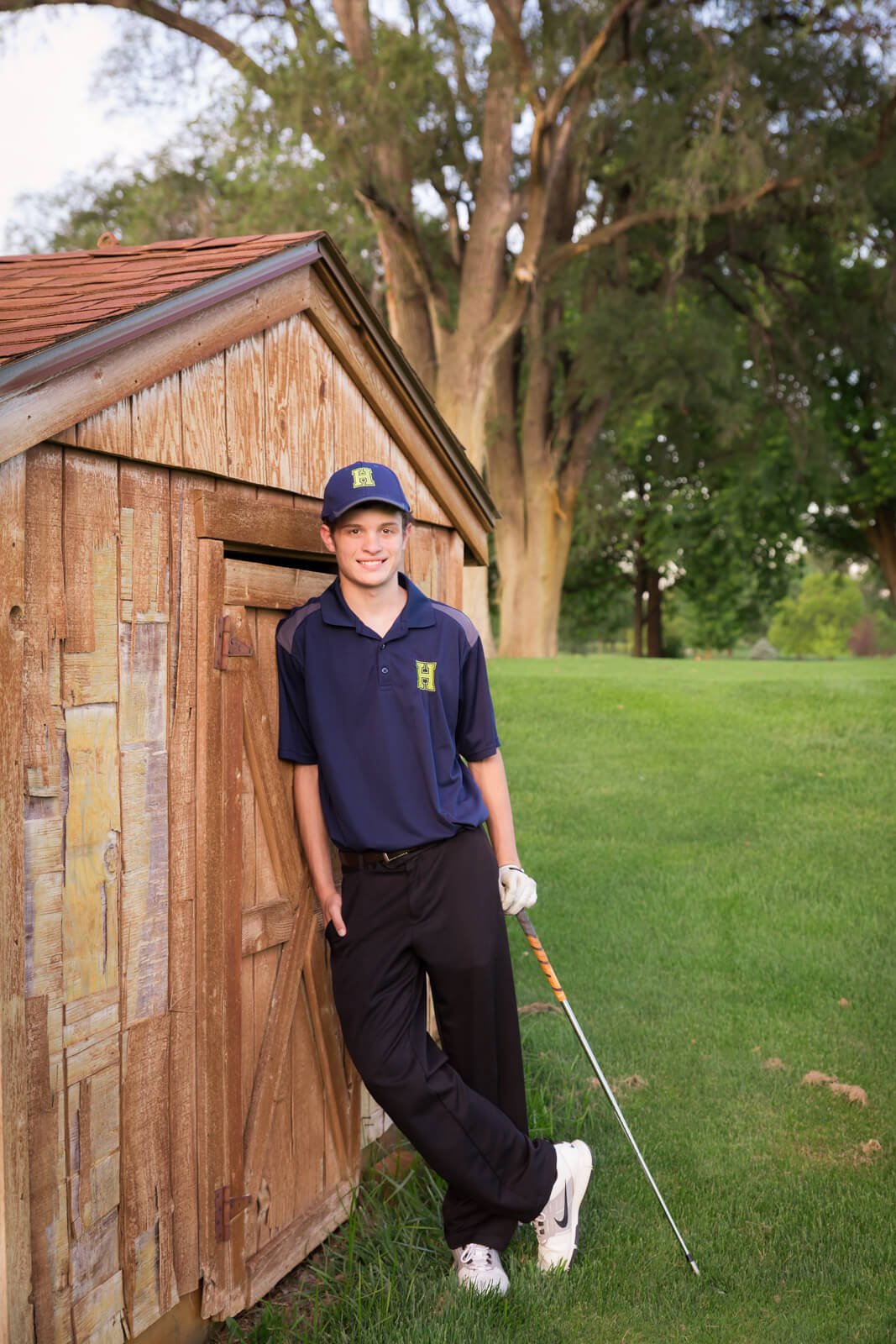 Senior boy leaning against a wooden shed, holding a golf club and wearing his school golf uniform.