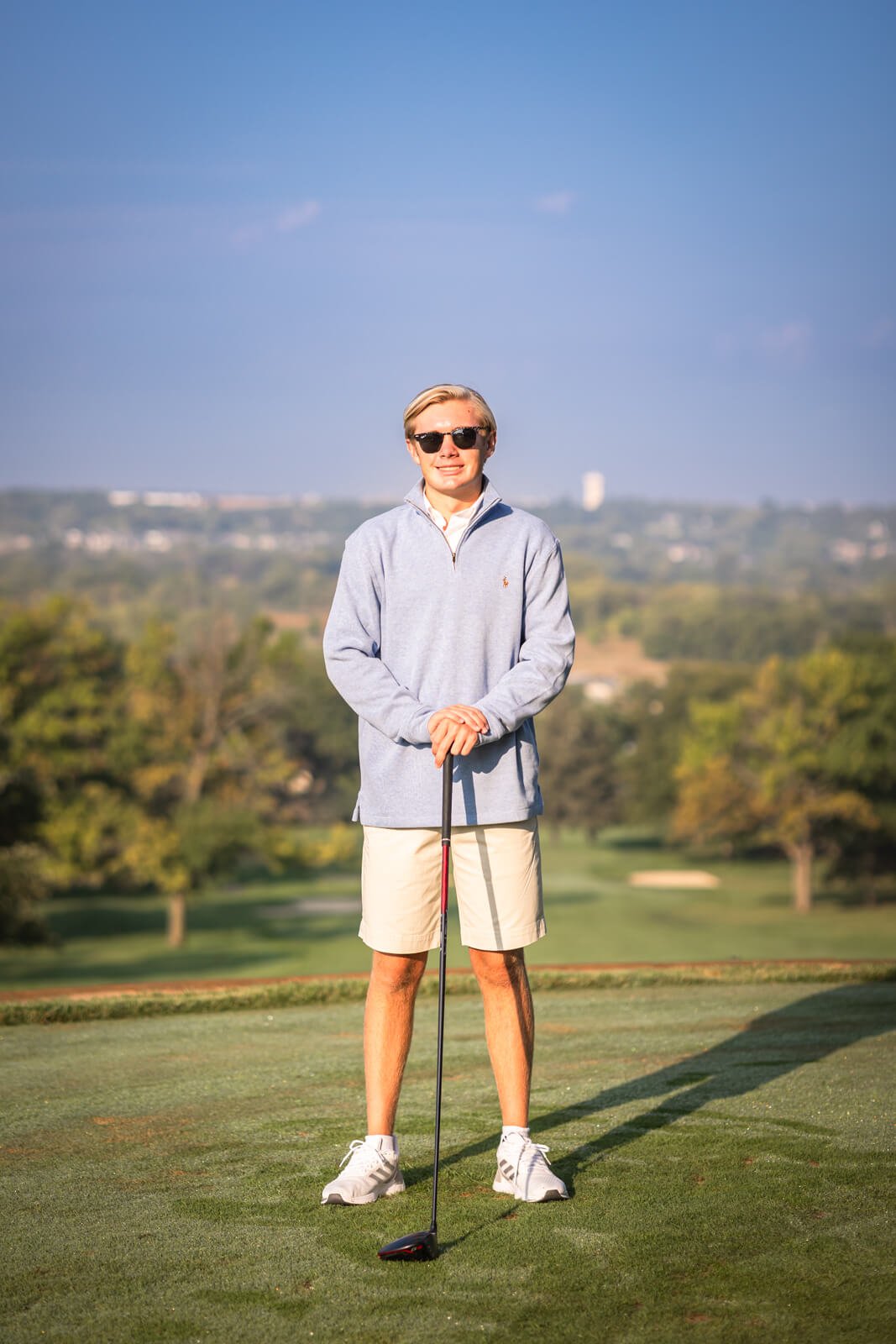 Senior boy wearing sunglasses and a blue half-zip sweater, standing on golf course while holding his golf driver.