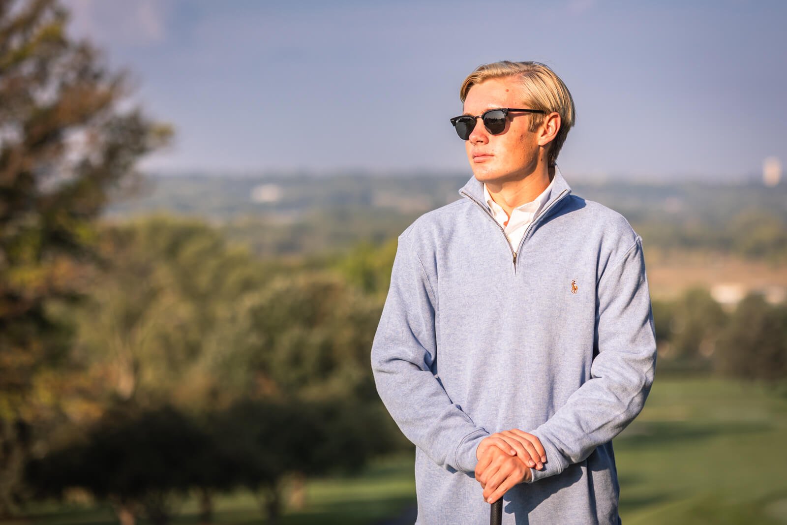 Blonde senior boy wearing blue half zip sweater, looking off to the side, wearing sunglasses, on a golf course in Johnston, Iowa.
