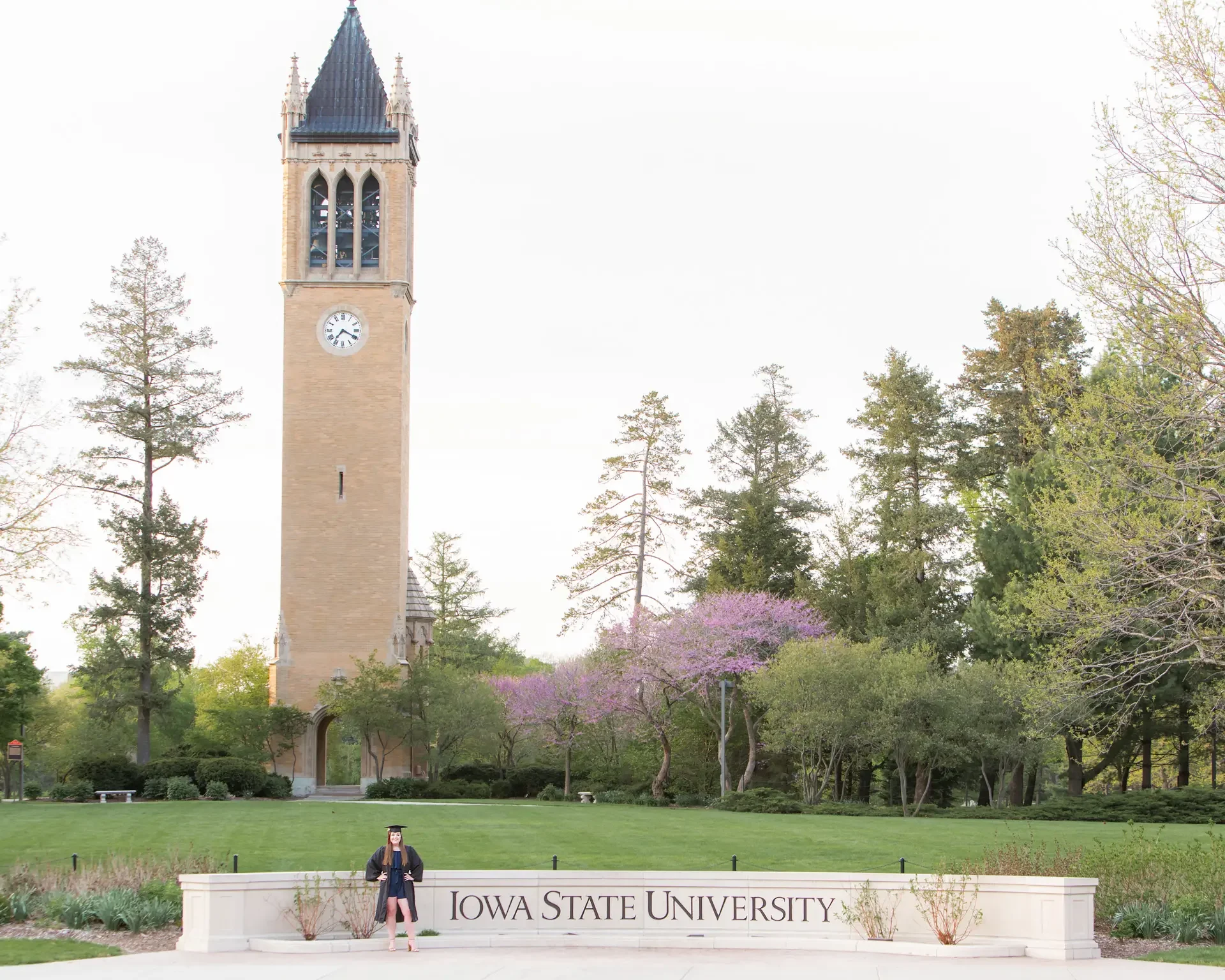 College grad wearing cap and gown in front of the Iowa State University sign, with the Campanile in the background, in Ames, Iowa.