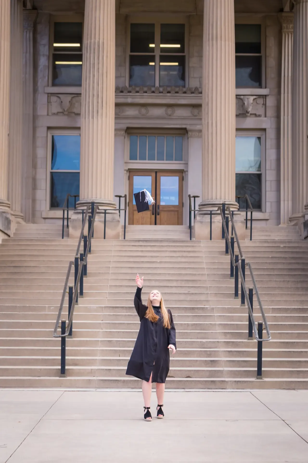 College grad tossing cap in the air in front of the steps of Curtis Hall at Iowa State University.