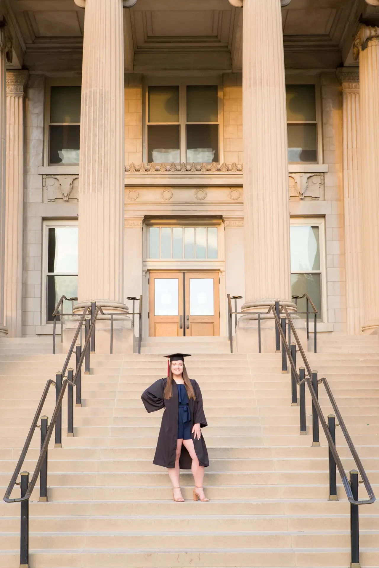 College graduate wearing cap and gown, standing with hand on hip, on the steps of Curtis Hall at Iowa State University.