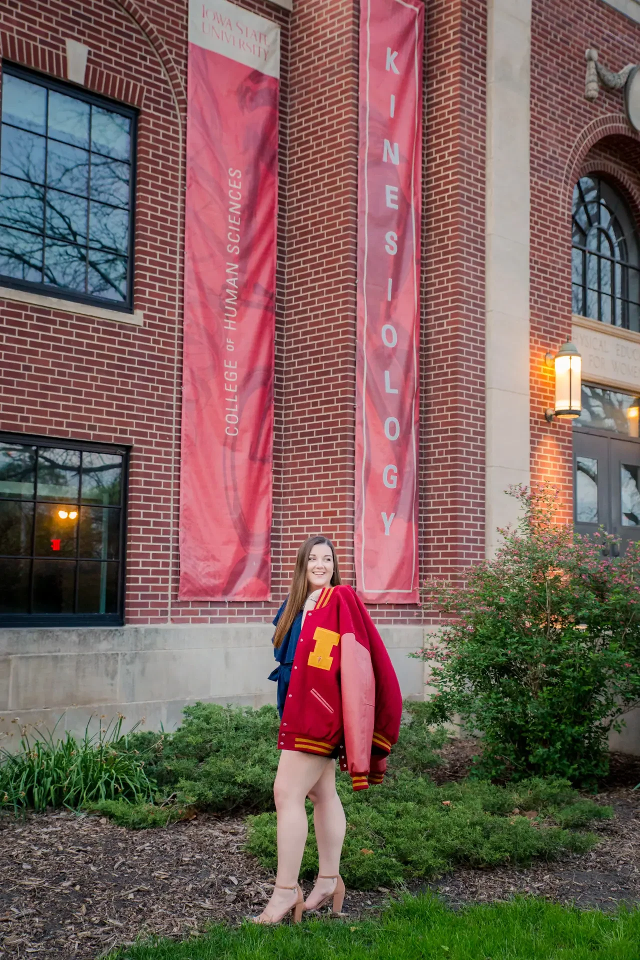 College grad holding her dad's ISU letter jacket in front of the College of Human Sciences Kinesiology banners.