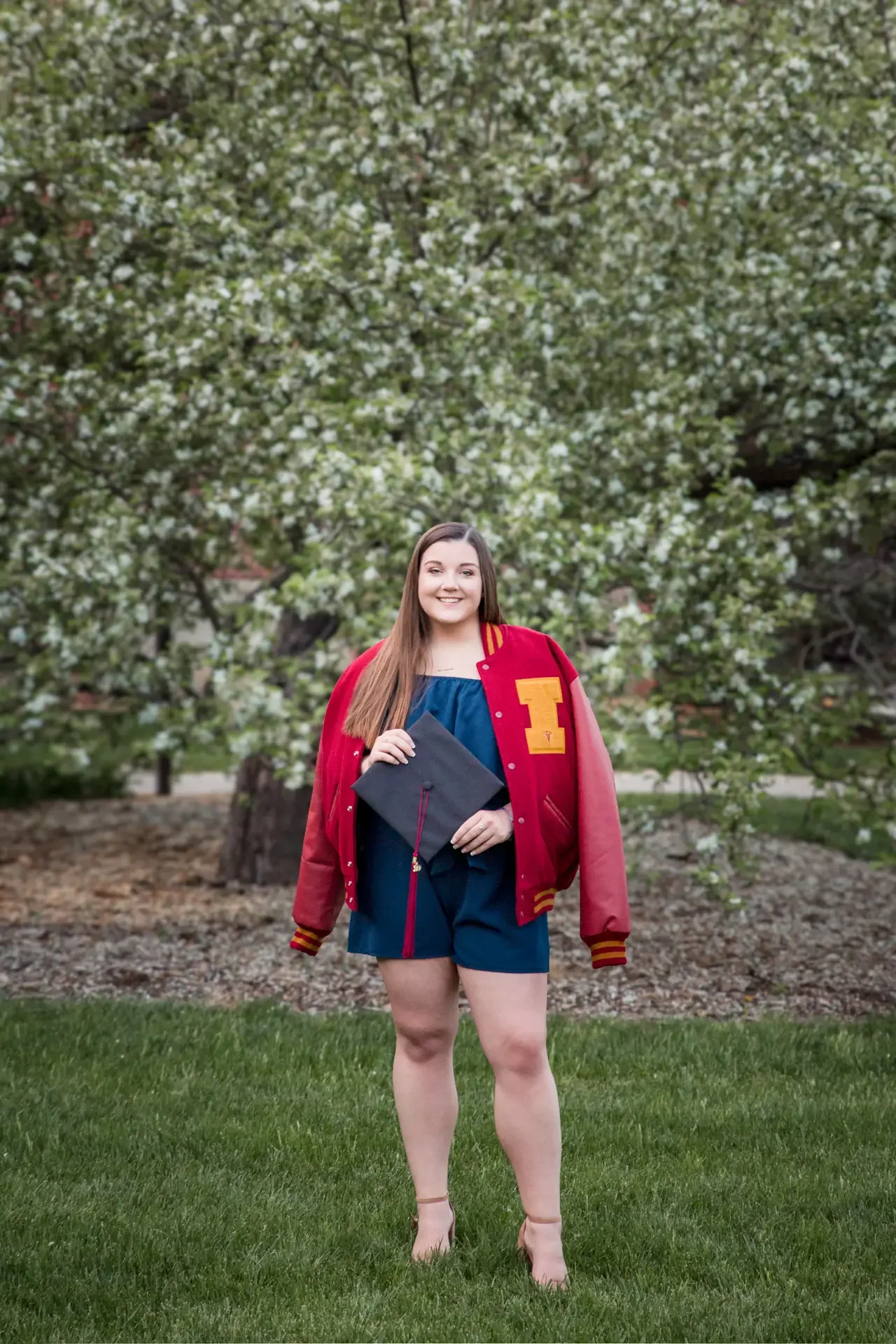 College senior wearing an ISU letter jacket, holding her graduation cap.