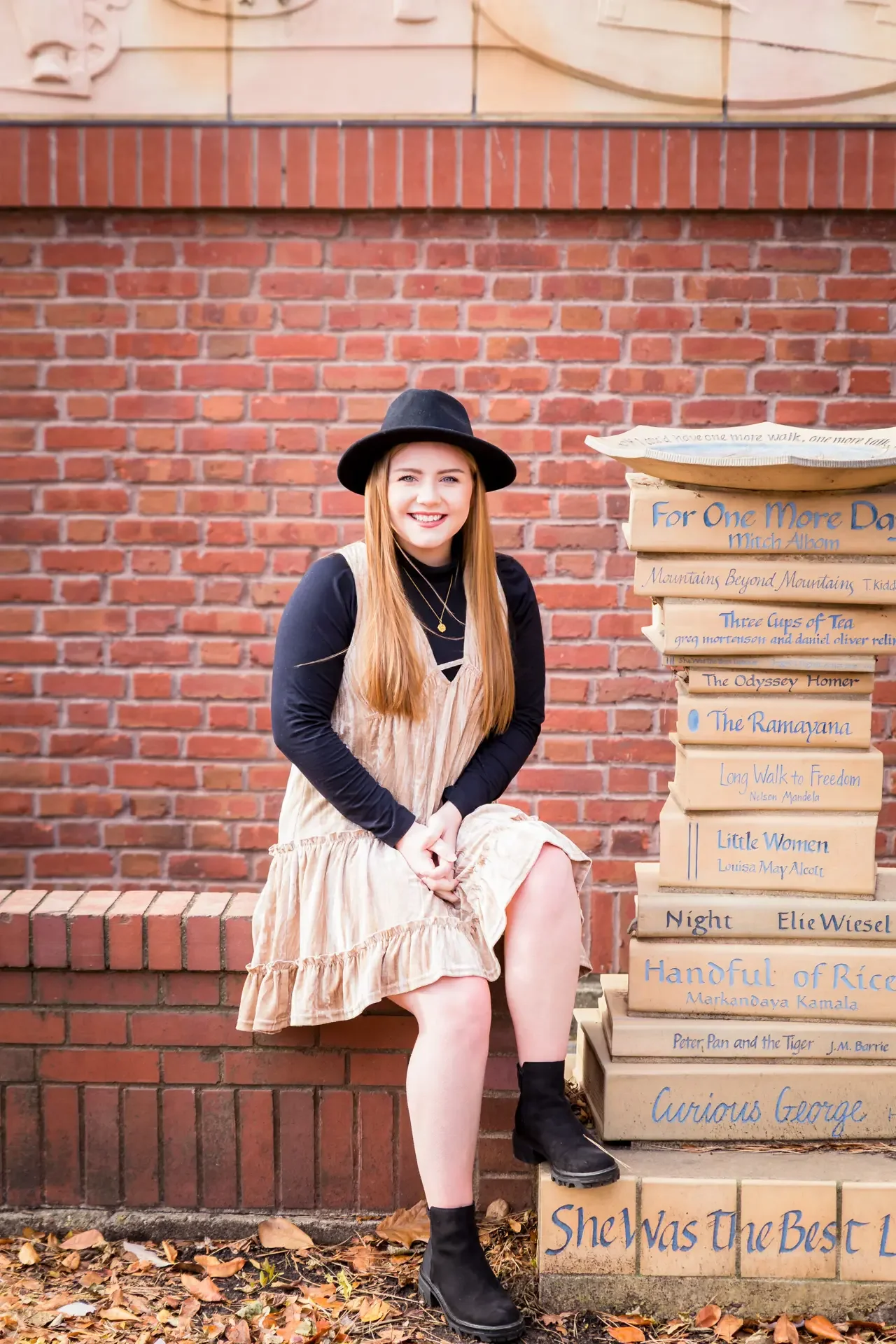 College girl wearing a dress and hat, sitting on a bench next to a sculpture of stacked books at Iowa State University.