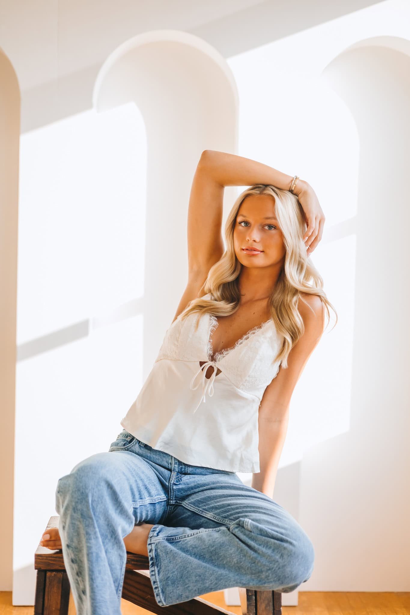 Senior girl with long blonde hair wearing jeans and a white lace trimmed top, sitting on a stool with her arm across the top of her head.