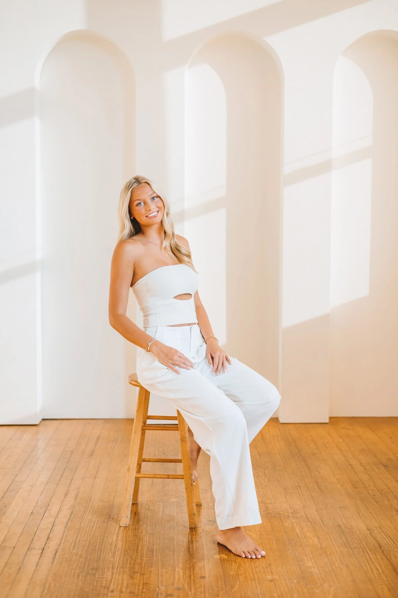 Senior girl wearing a white strapless pant suit, sitting on a wood stool, smiling at the camera.
