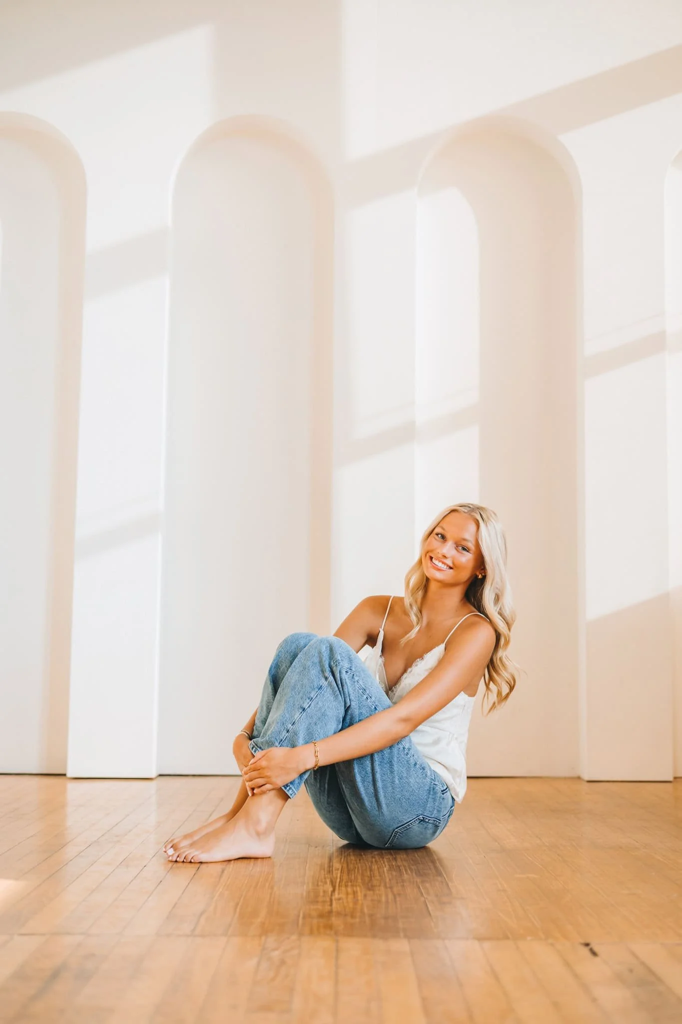 Senior girl with long blonde hair sitting on the floor with both knees bent, arms wrapped around her legs, smiling at camera.