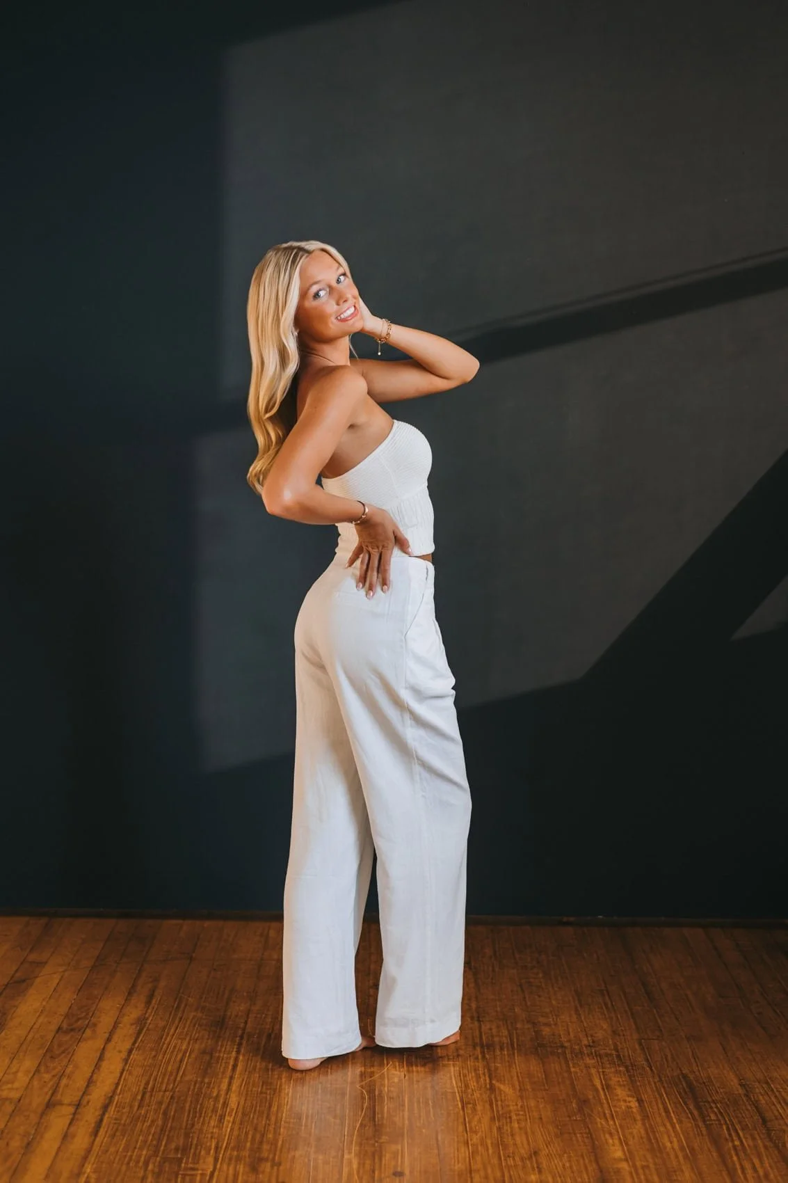 Senior girl with long blonde hair wearing a white strapless top and wide-leg white pants standing on a wooden floor against a dark wall.