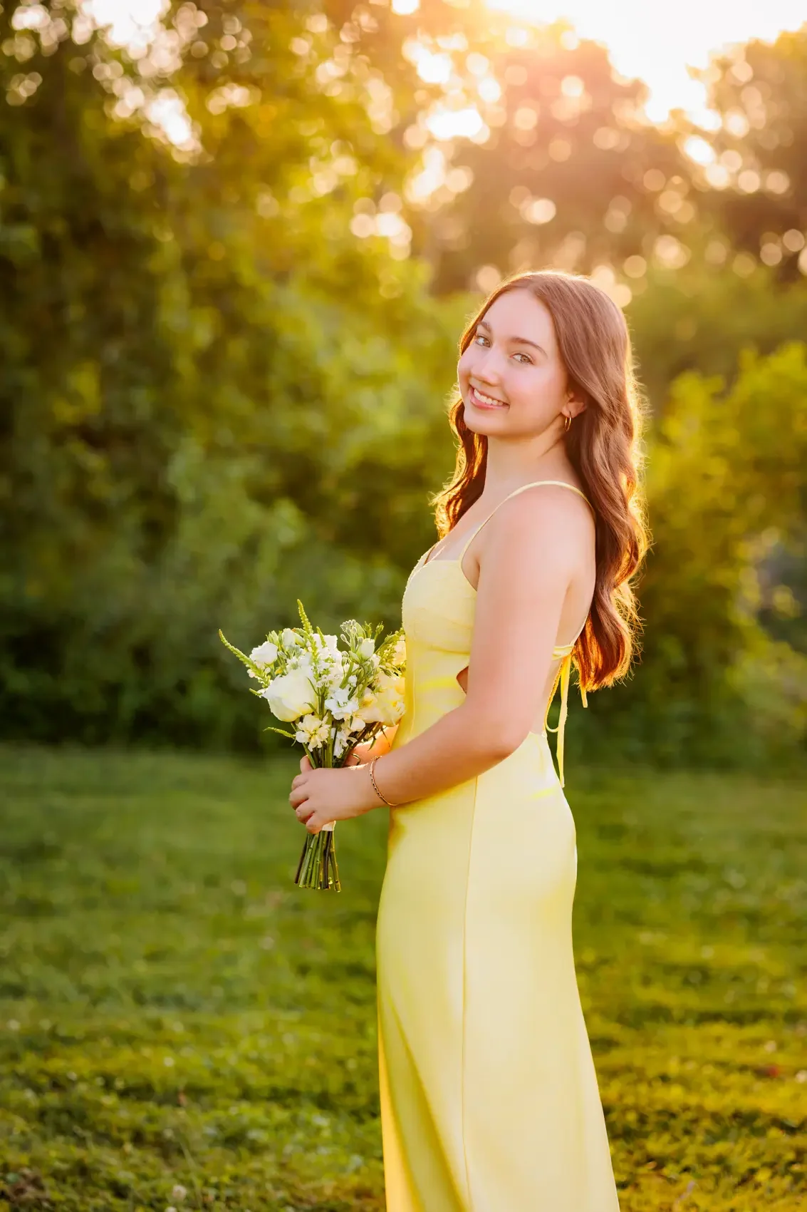 Senior girl in a yellow dress holding a bouquet of flowers outdoors during sunset, smiling at the camera. Photo by Des Moines senior photographer Wendy Sorensen.