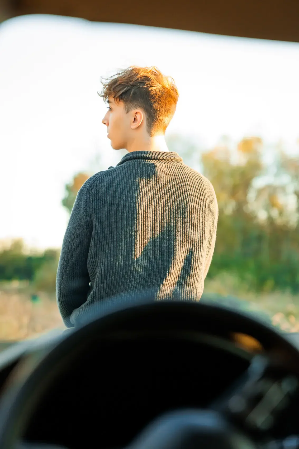 Young man with short, styled hair in profile facing left, standing outdoors during golden hour, wearing a dark ribbed sweater, with blurry trees in the background, seen from inside a vehicle. Photo by Wendy Sorensen, a Des Moines senior photographer.
