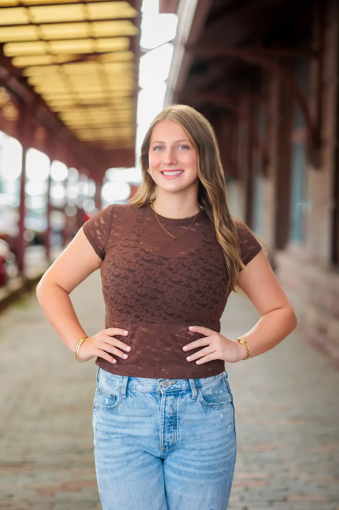 High school senior girl with brown hair smiling, standing with hands on hips, outside under a canopy in Des Moines with a blurred background.