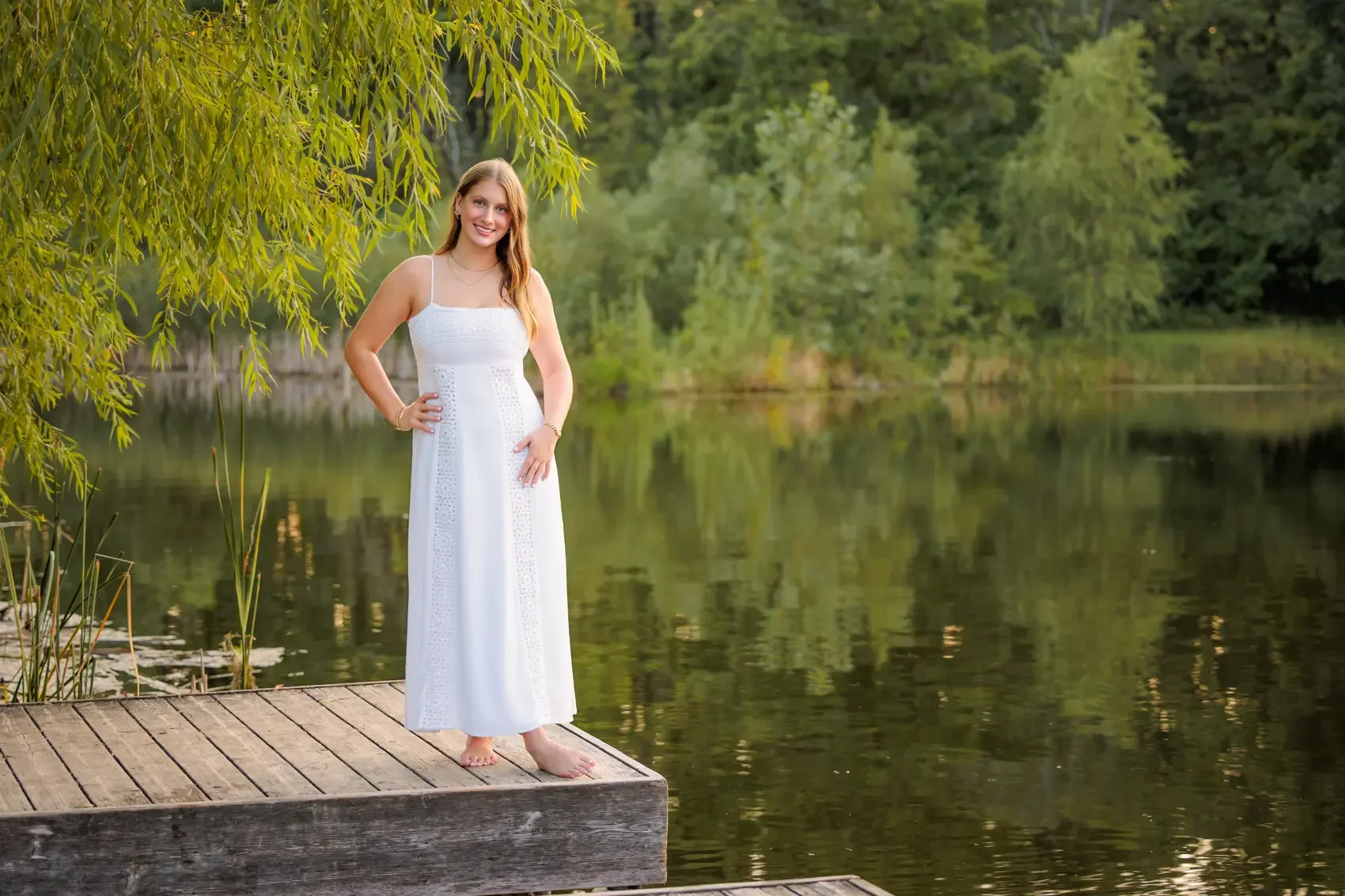 Senior girl  in a white dress standing barefoot on a wooden dock by a lake, surrounded by green trees and foliage.