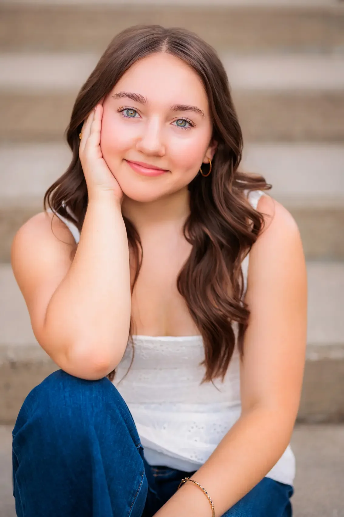 A young woman with long, wavy brown hair and blue eyes smiling softly, resting her hand on her cheek, sitting outdoors on cement steps.