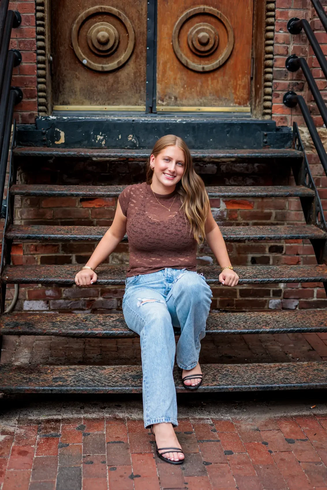 Senior girl with long blonde hair sitting on outdoor brick stairs in front of a large wooden door, smiling and wearing a brown lace top, ripped light blue jeans, and black strappy high-heeled sandals.  