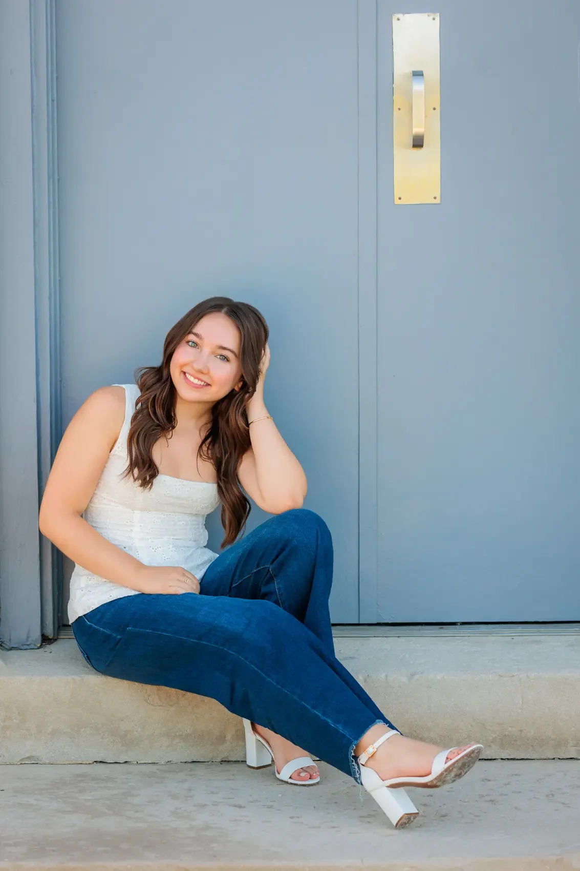 Young woman with long brown hair, smiling, sitting on a sidewalk with her back against a blue door, wearing white heeled sandals, a white sleeveless top, and blue jeans.