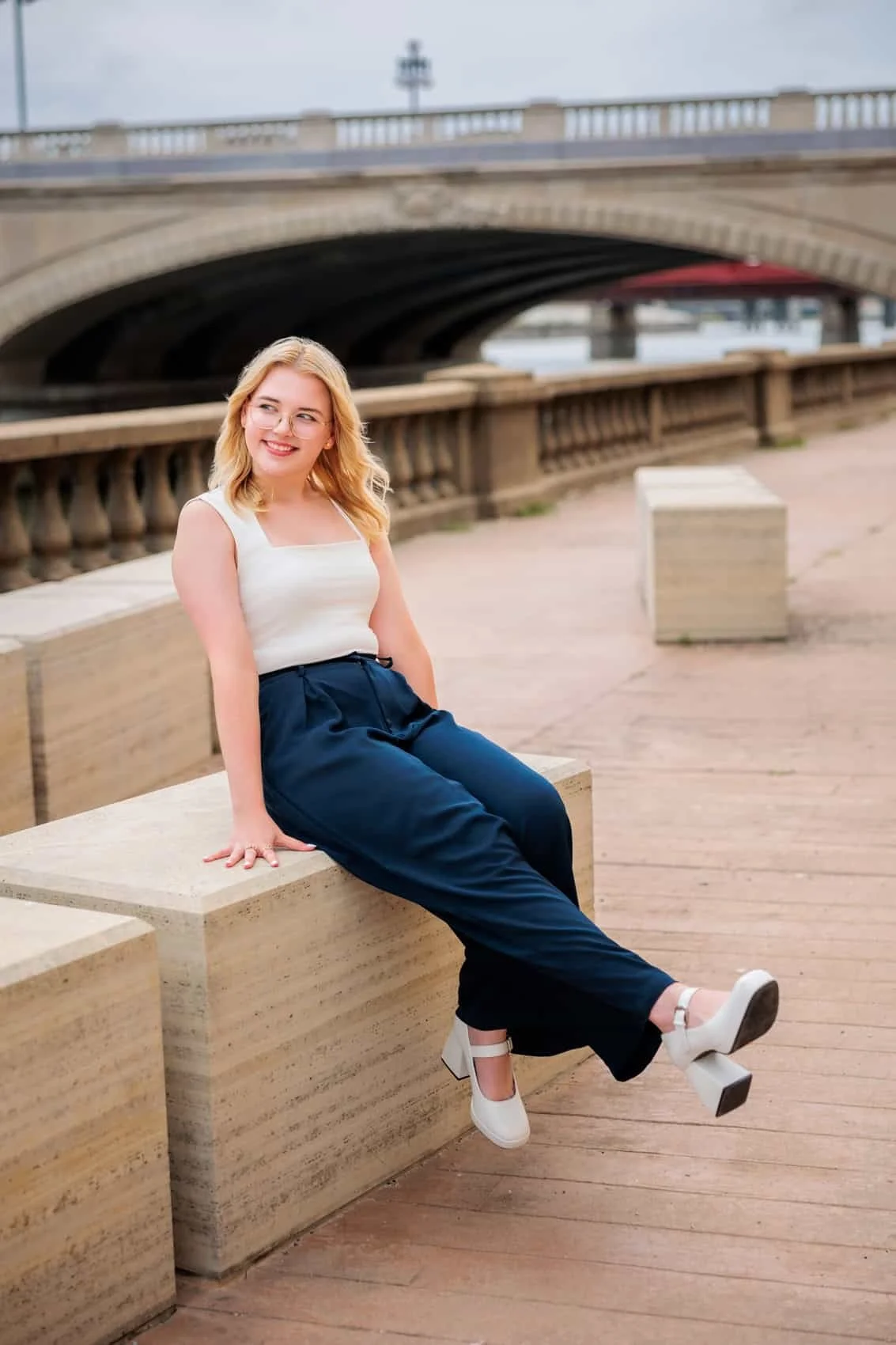  High school senior girl with blonde hair, glasses, wearing a white sleeveless top, navy blue high-waisted pants, and white heels, sitting on a concrete bench near a bridge by the Des Moines River.