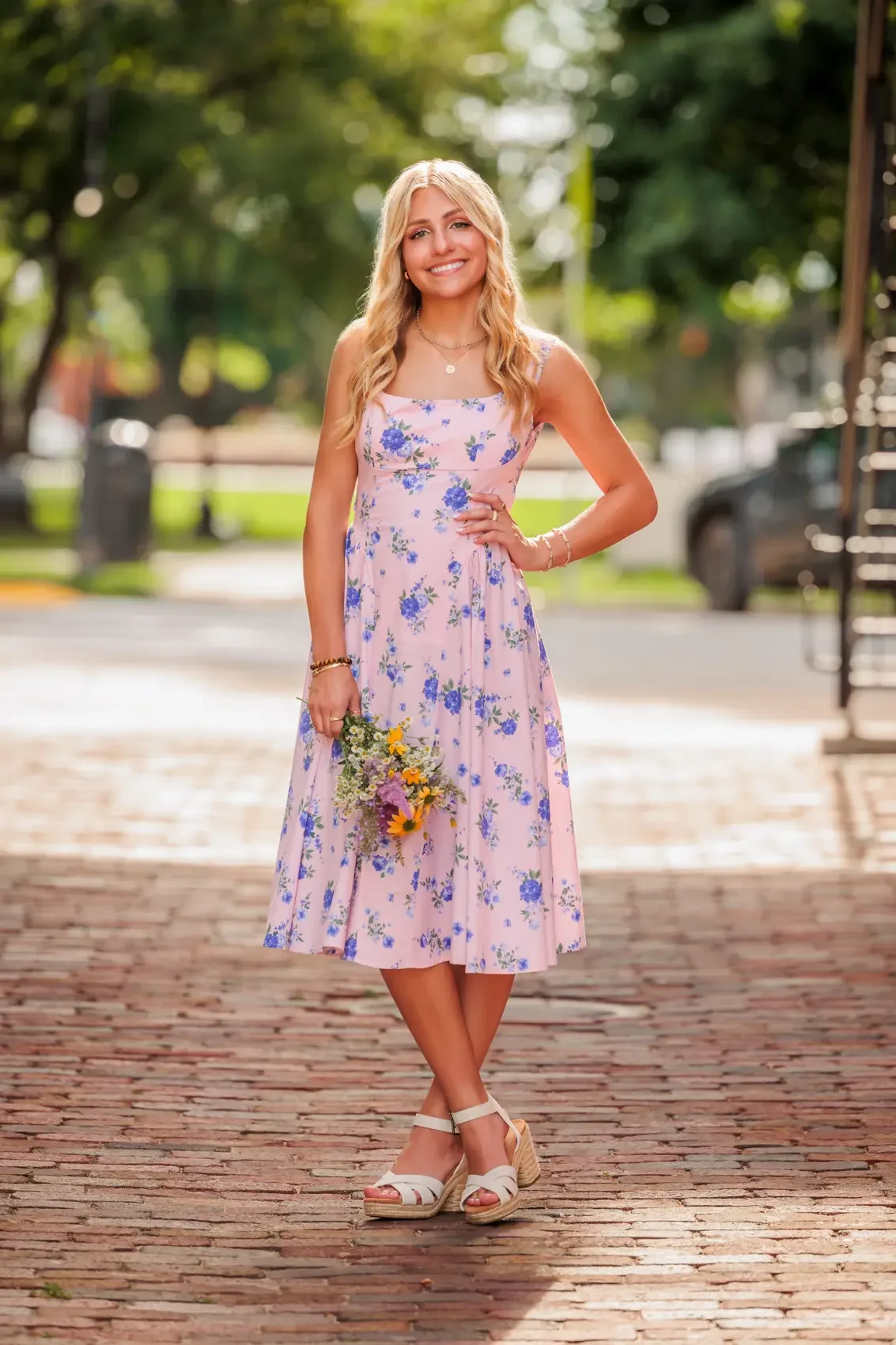 A young woman with blonde hair wearing a pink floral dress holding a small bouquet of flowers standing outdoors on a brick pathway.